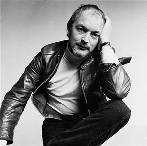 Black and white photo of a man with a beard and receding hairline, wearing a leather jacket, sitting with one leg crossed, resting his head on one hand.