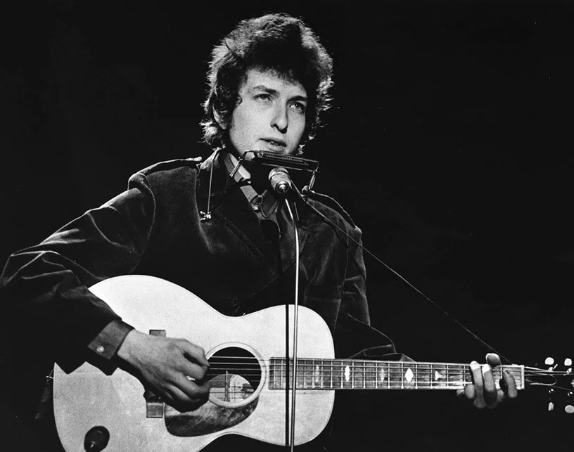 A black-and-white photo of a young man with curly hair playing an acoustic guitar and singing into a microphone on a stage.