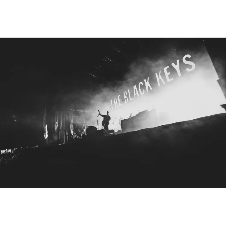 A black and white photo of a live concert by The Black Keys, with a band performing on stage and a large sign displaying their name in the background.