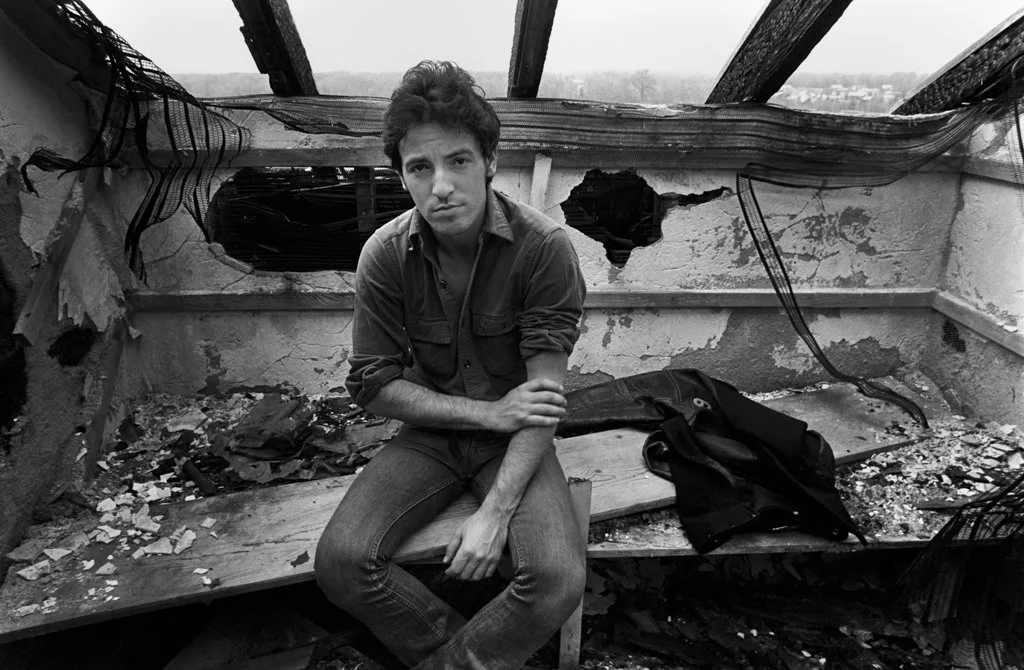 A young man with curly hair and a concerned expression, sitting on a bench in a damaged, abandoned building with holes and debris.