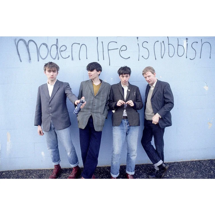 Four young men standing against a light blue wall with graffiti that reads "modern life is rubbish."