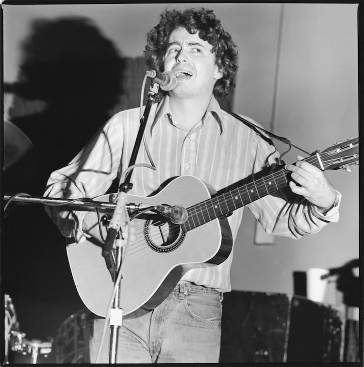 A man with curly hair singing into a microphone while playing an acoustic guitar.