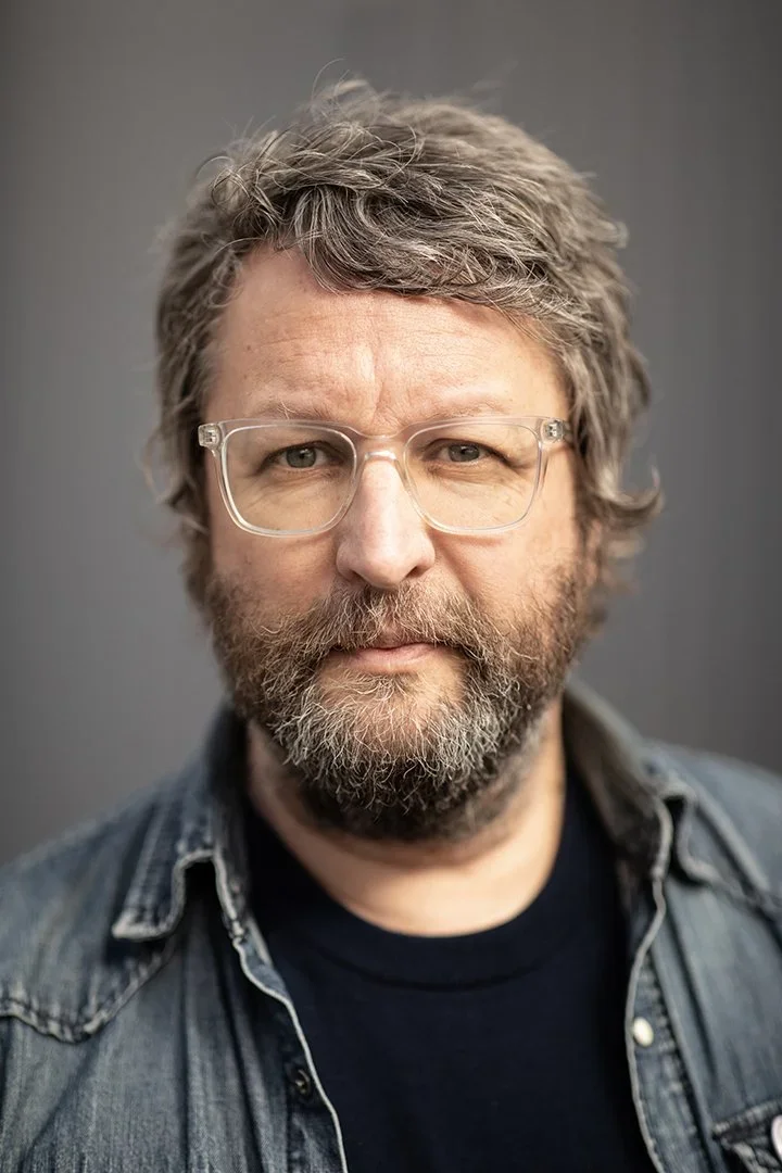 A middle-aged man with glasses, a full beard, and gray hair, wearing a black shirt and a denim jacket, standing against a gray background.