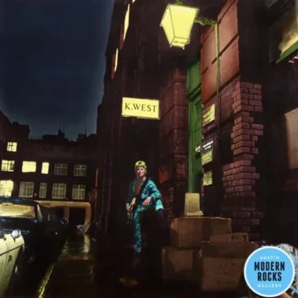 A person standing under a yellow street lamp and a sign that reads "K. West" on a city street at night. Buildings and parked cars are visible in the background. A blue circular sign in the bottom right corner says "Austin Modern Rocks Gallery."