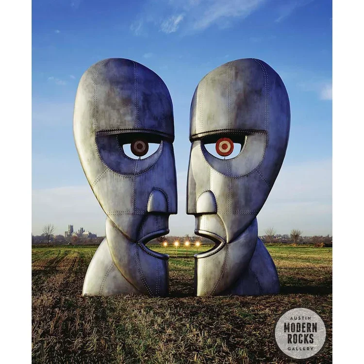 Large metal sculpture of two stylized faces close together facing each other, with one eye on each face having a target-like design, set in an open field under a blue sky.