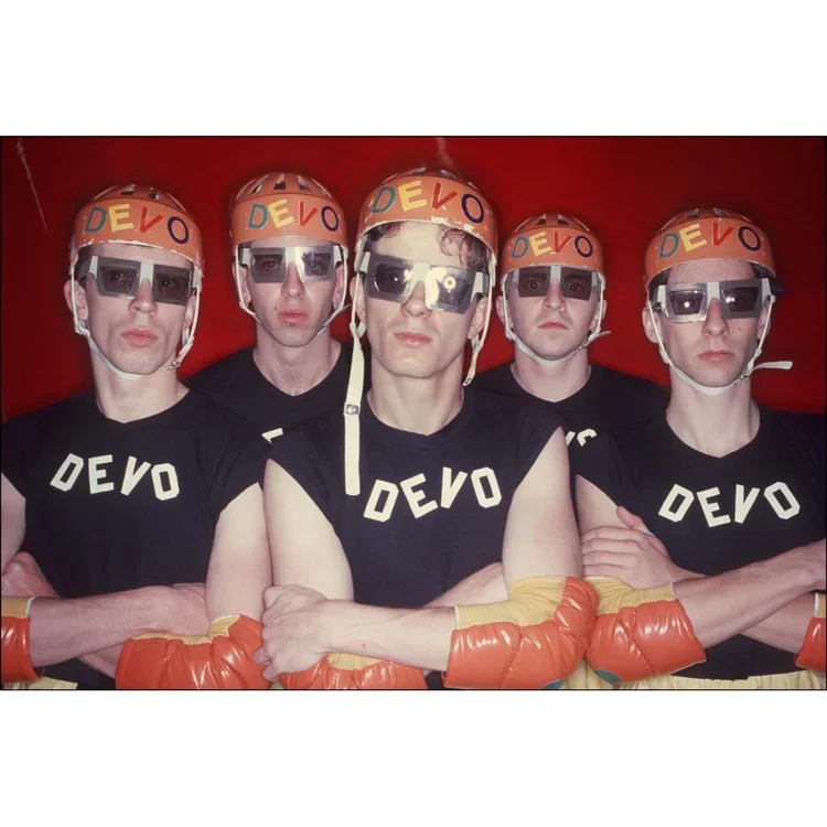 Five men dressed as boxers wearing referee helmets with 'DEVO' written on them, black 'DEVO' shirts, sunglasses, and orange boxing gloves, standing against a red background with arms crossed.
