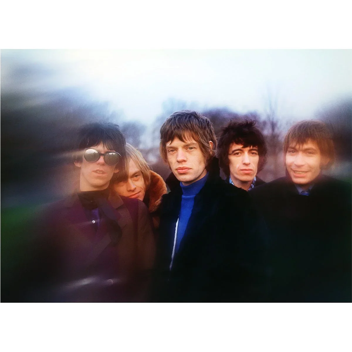 A color photograph of five young men standing outdoors with an out-of-focus background, some wearing dark clothing and sunglasses, with serious or neutral expressions.