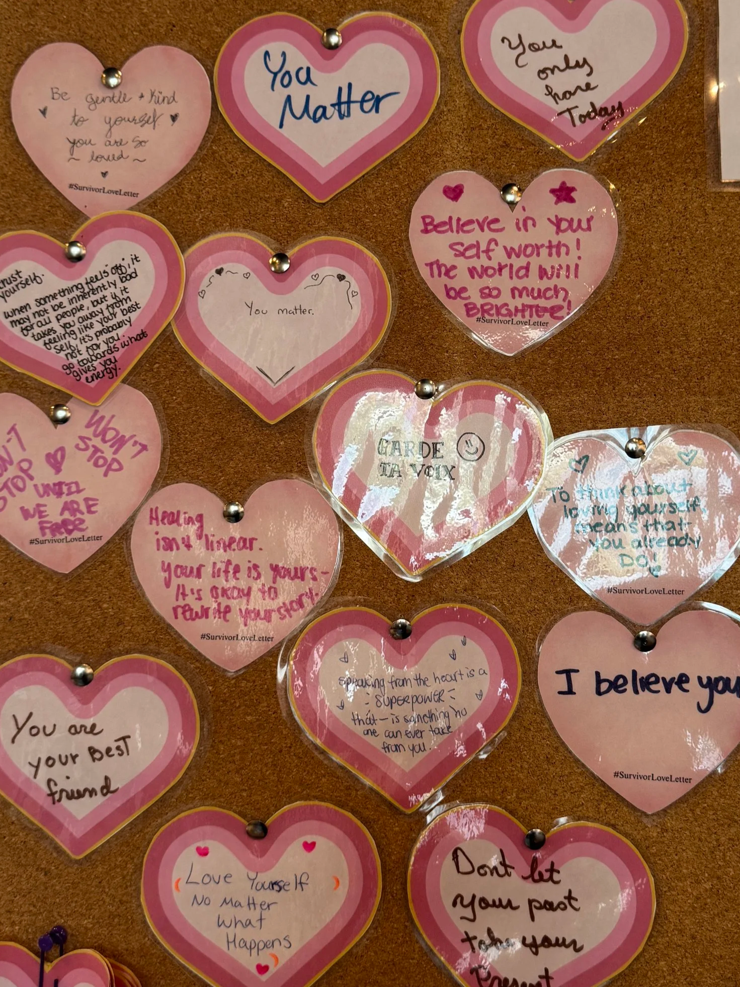 A bulletin board decorated with pink and transparent heart-shaped notes containing positive affirmations and motivational messages about self-love, kindness, and confidence.