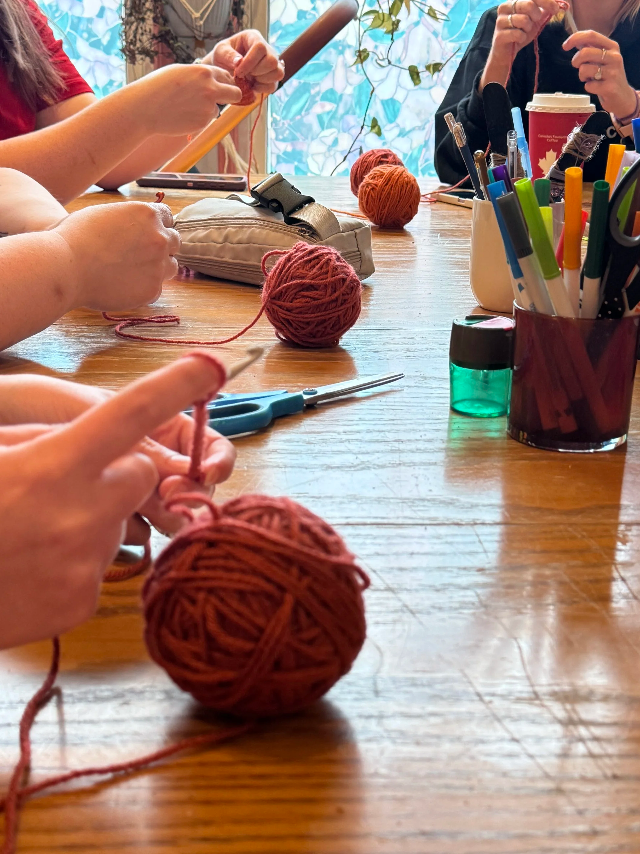 People knitting with balls of yarn, scissors, and knitting supplies on a wooden table inside a room.