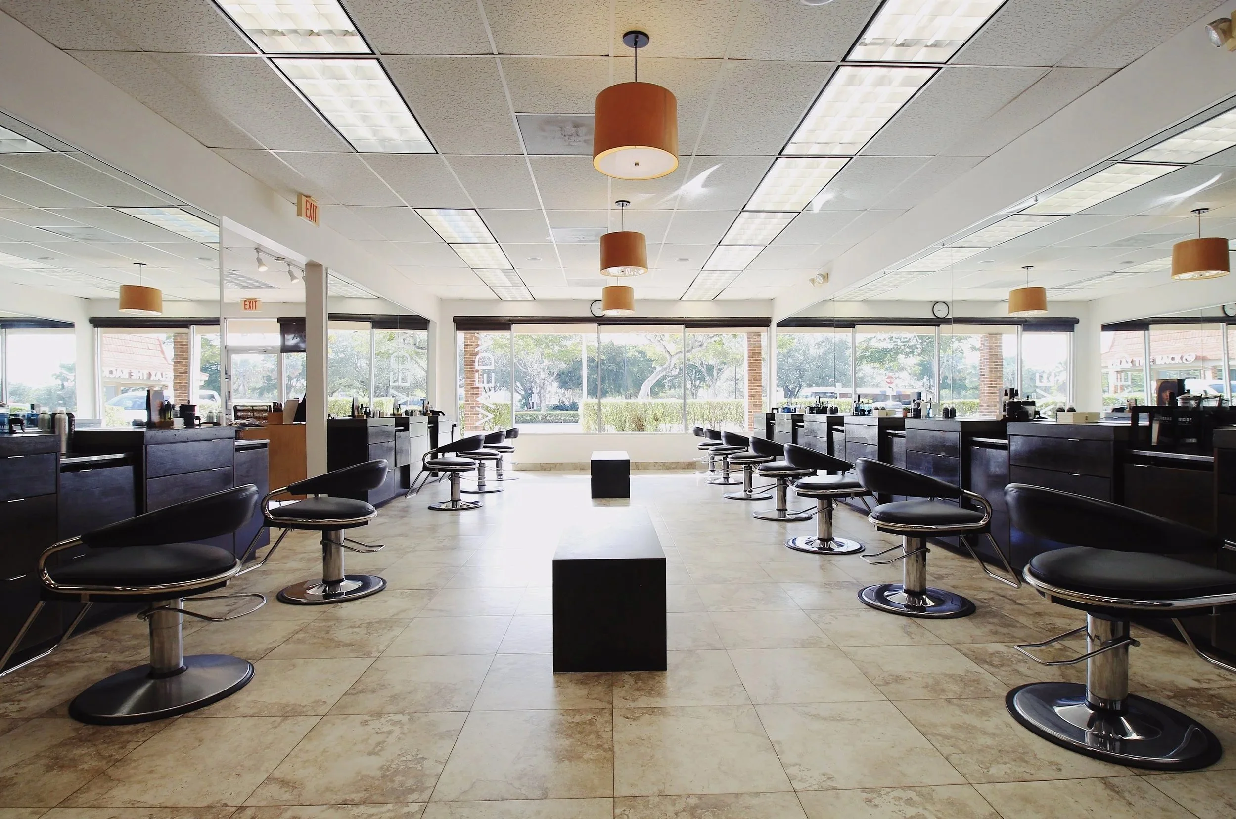 Empty hair salon with black chairs, mirrors, and large windows showing outside view.
