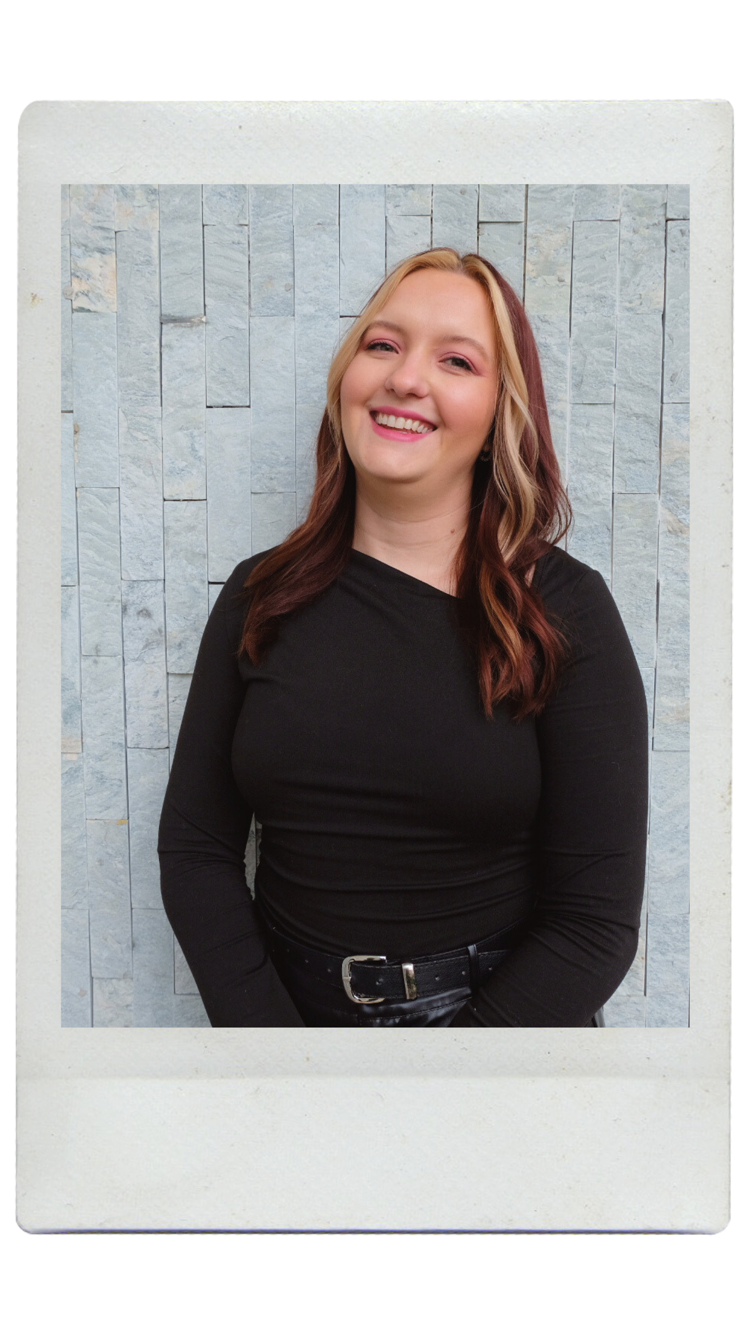 A woman with blonde and brown hair smiling, wearing a black long-sleeve shirt, standing against a textured light-colored stone wall.