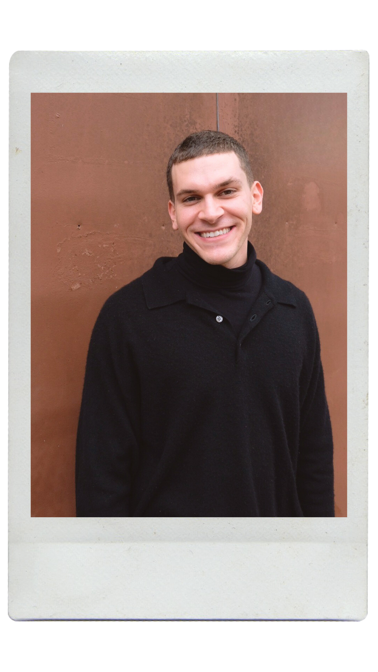 A young person with curly hair smiling at the camera, wearing a black shirt and layered necklaces, standing against a textured wall.