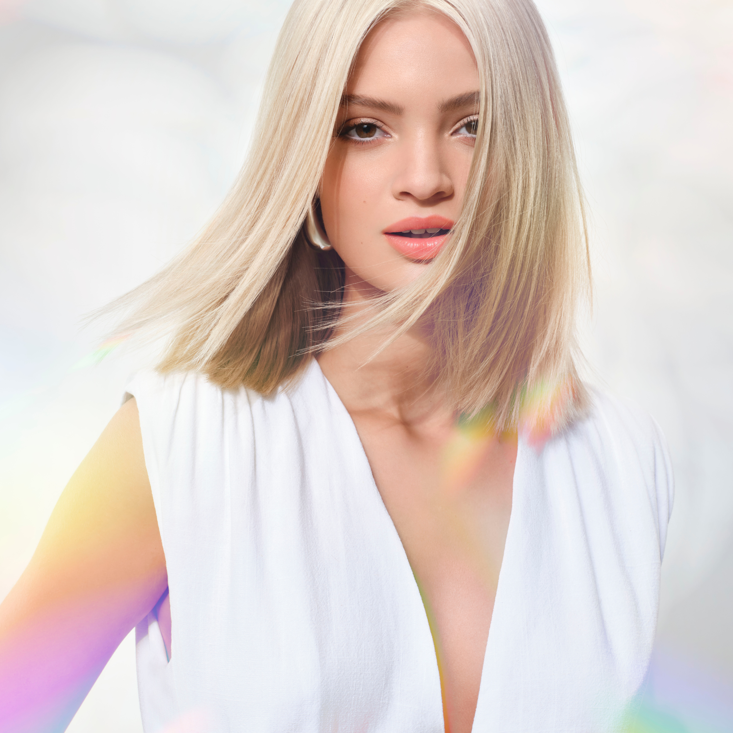 A young woman with blonde hair, wearing a white sleeveless top, looking into the camera against a light background with rainbow reflections.