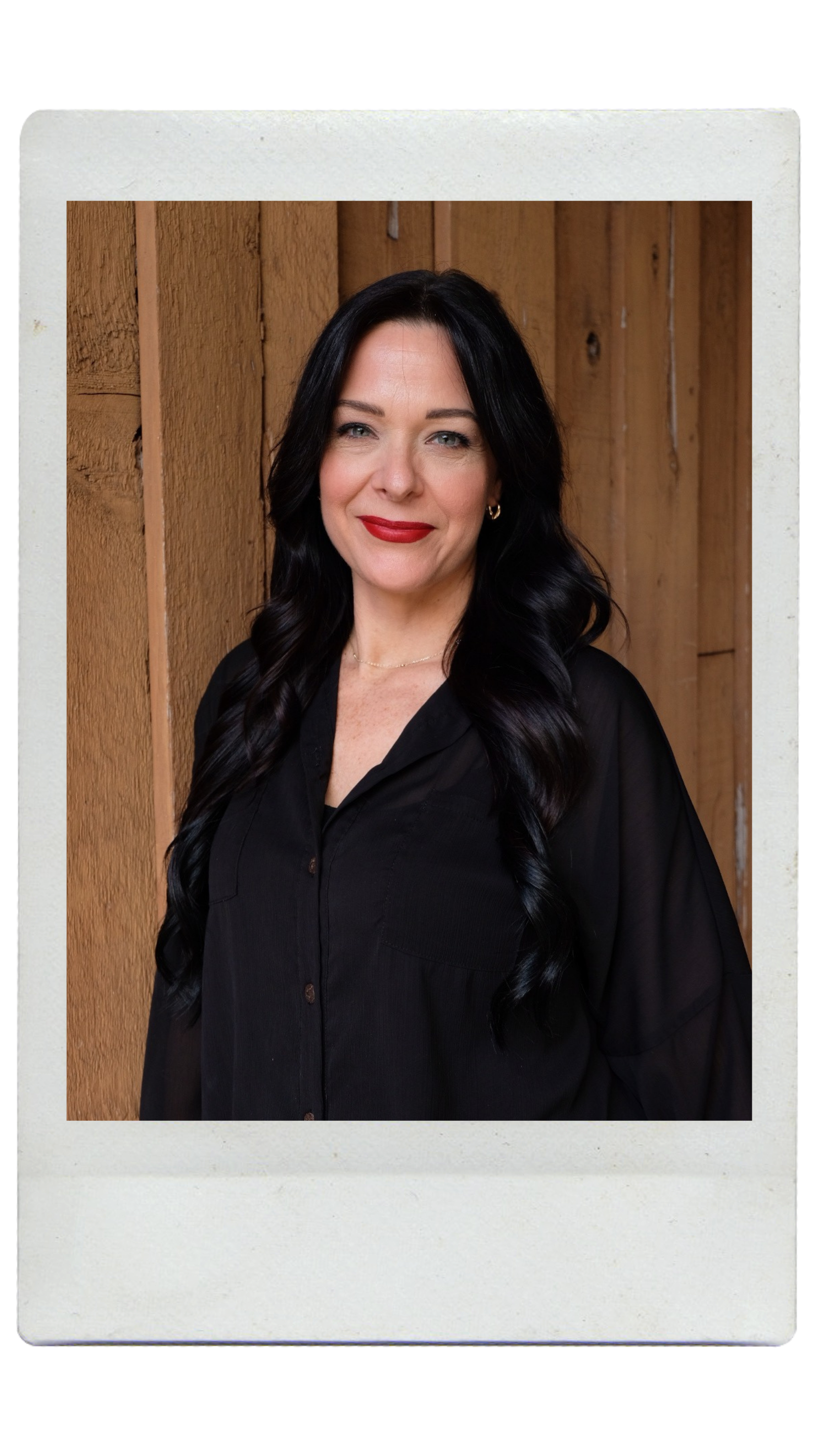 A woman with long brown hair and blue eyes smiling, wearing red lipstick, a black top, and a gold necklace with a rectangular pendant, in front of a plain light-colored background.