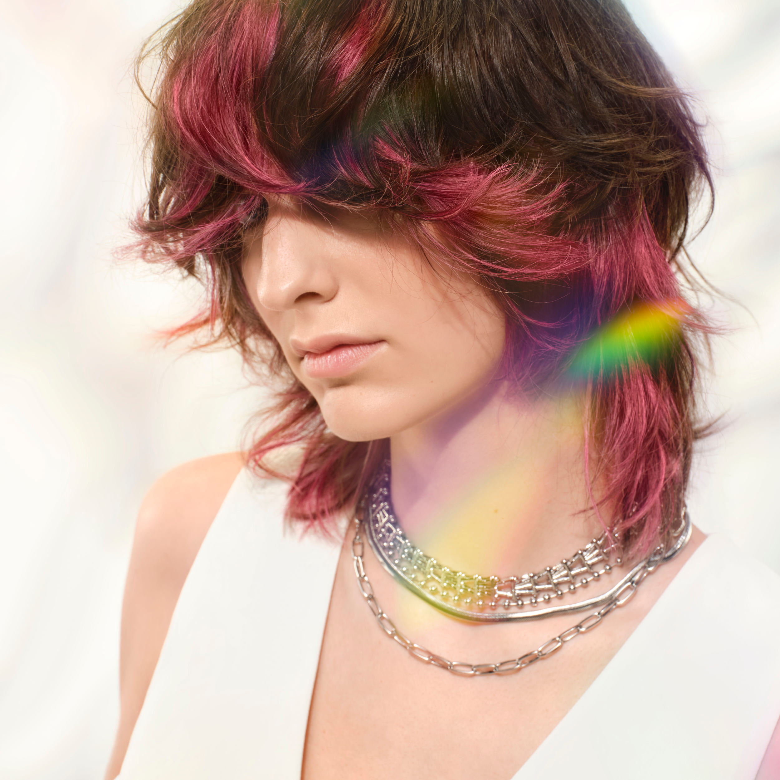 A woman with multicolored pink and purple hair looking down, wearing layered silver necklaces, and dressed in a white sleeveless top, with soft lighting and a blurred white background.