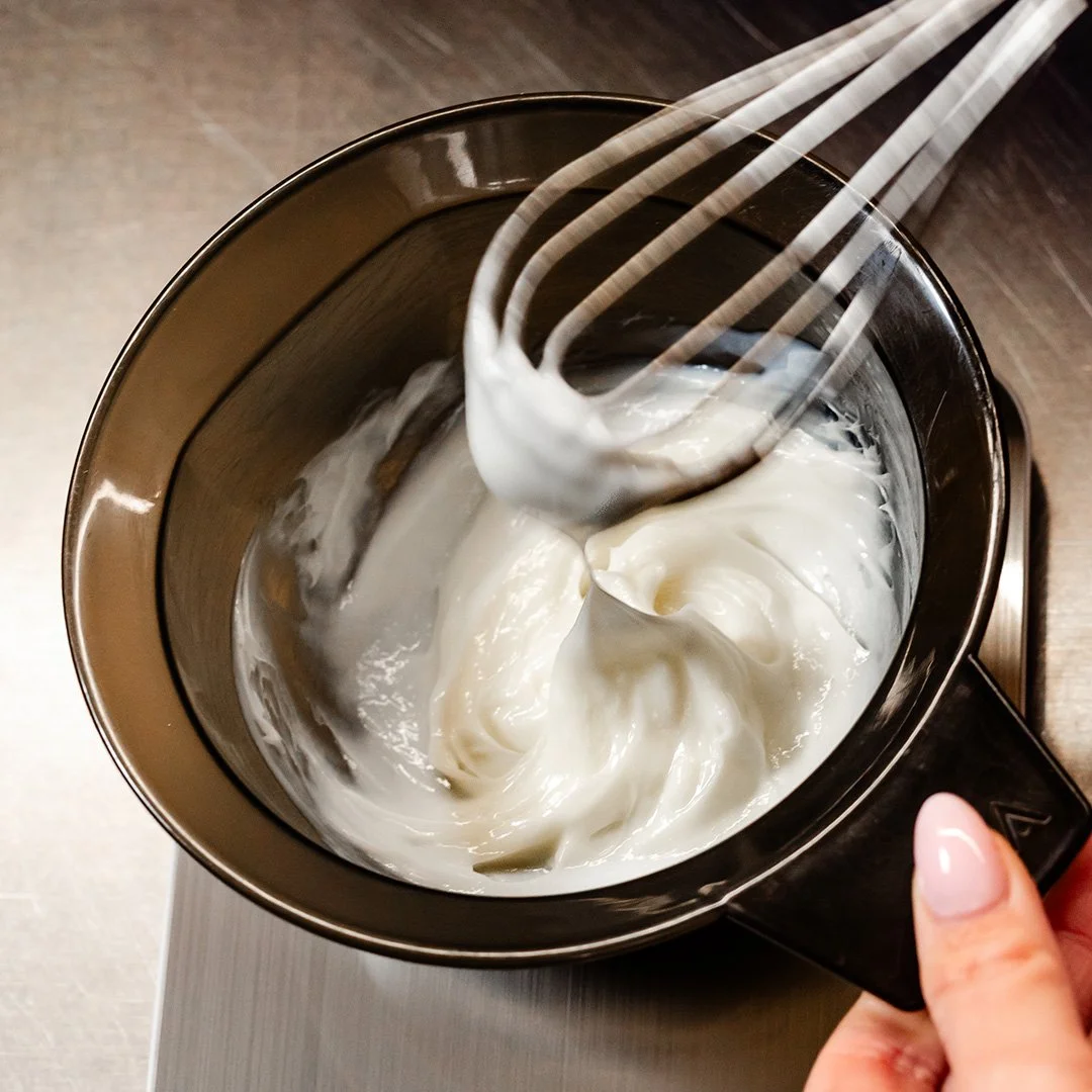 A hand holding a small metal container with white whipped cream, a whisk is being dipped into it.