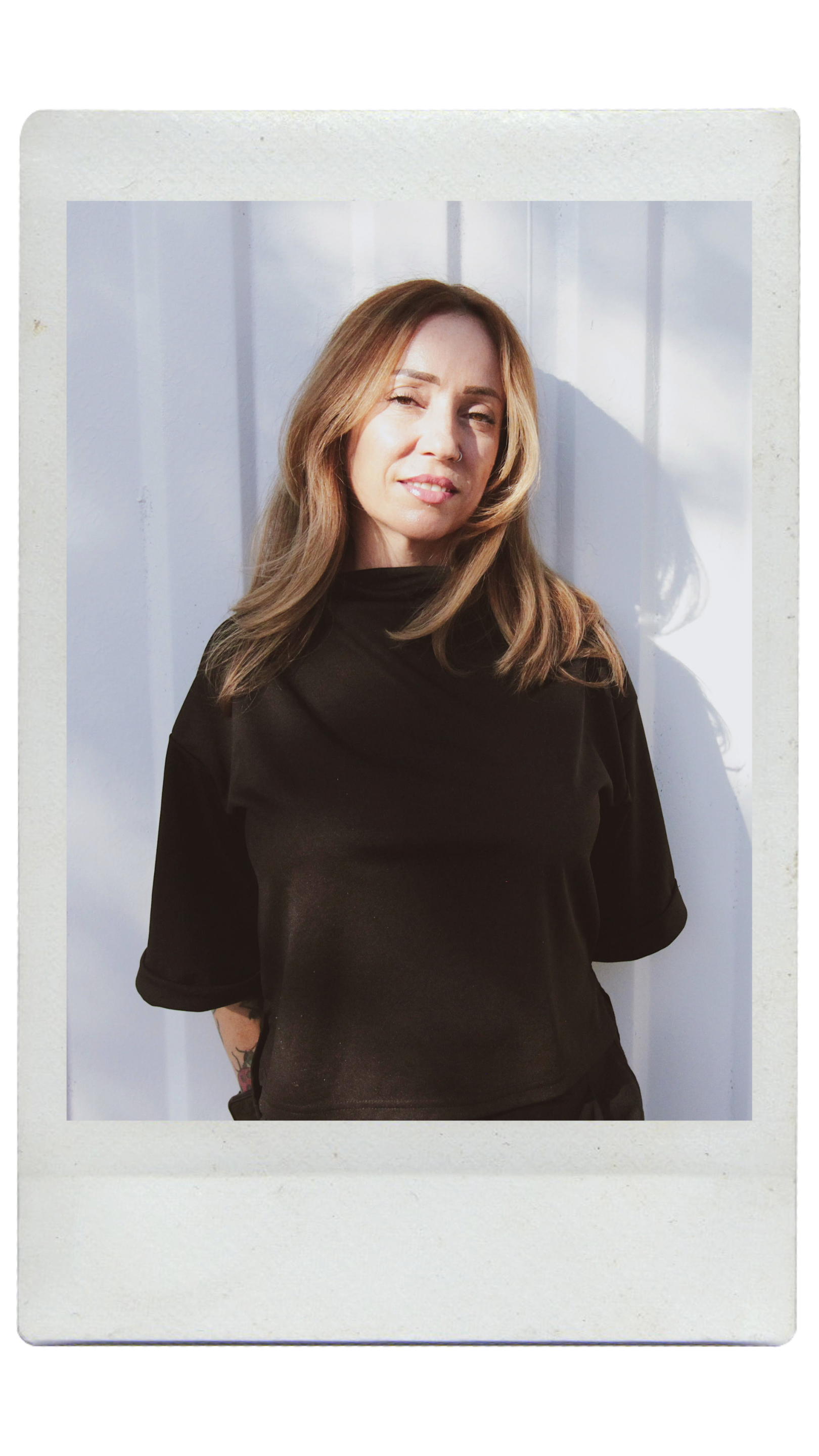 A woman with long, light brown hair, wearing a black top, standing outdoors in bright sunlight, with a white wall background.