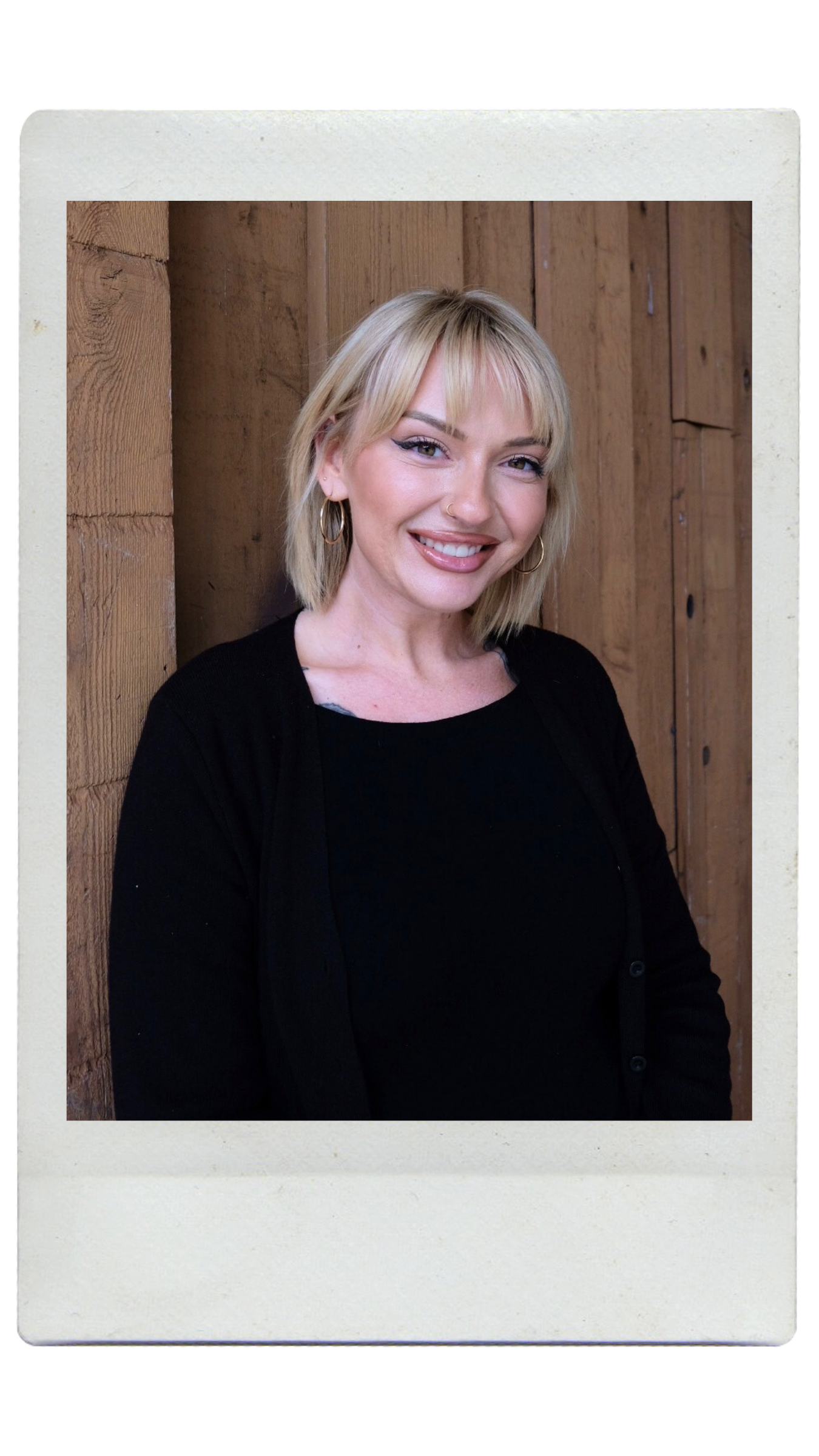 A woman with blonde hair styled in a bob, wearing a black long-sleeve top, standing against a light-colored textured wall, smiling at the camera.