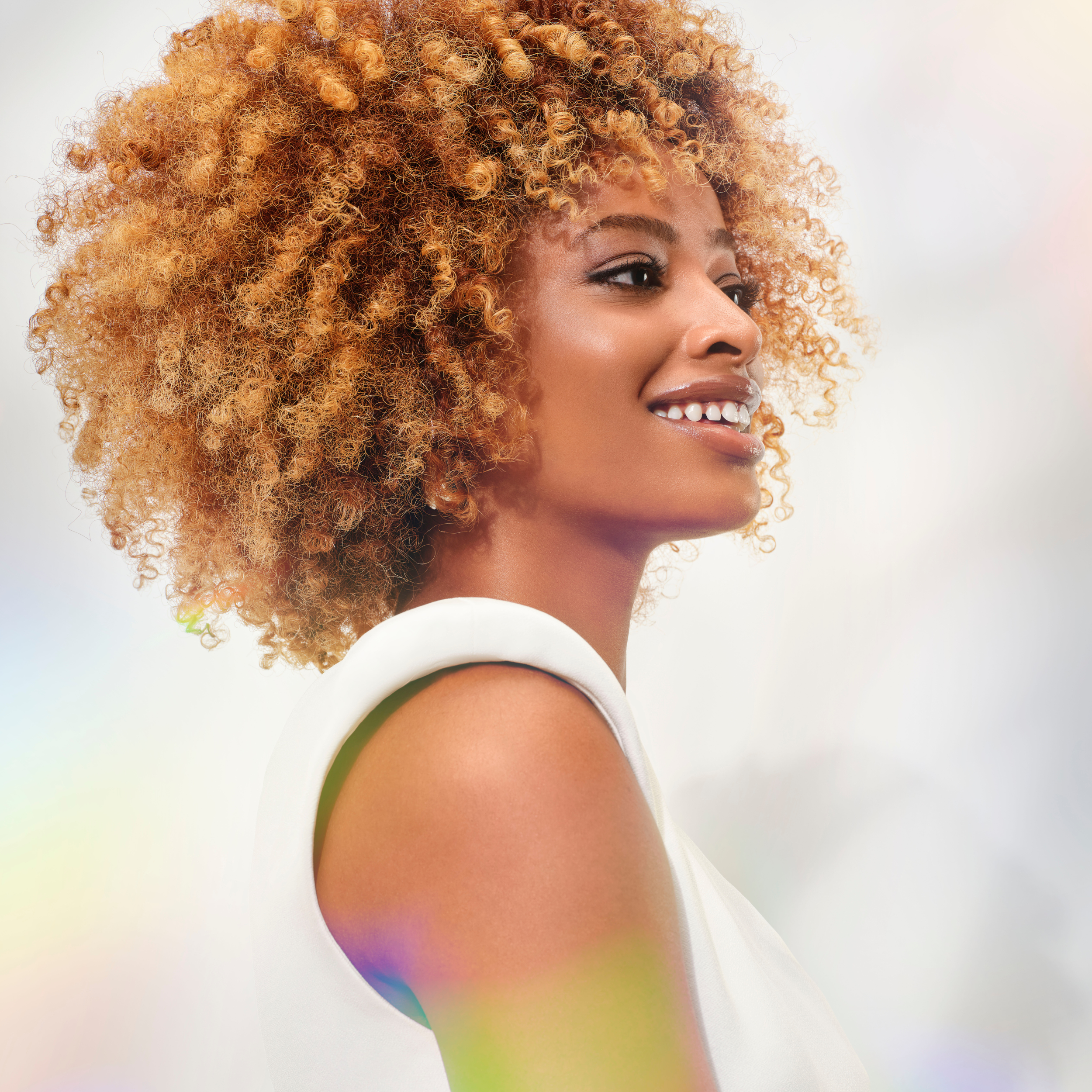 Side profile of a smiling young woman with curly blonde hair, wearing a white sleeveless top, against a soft, out-of-focus background.