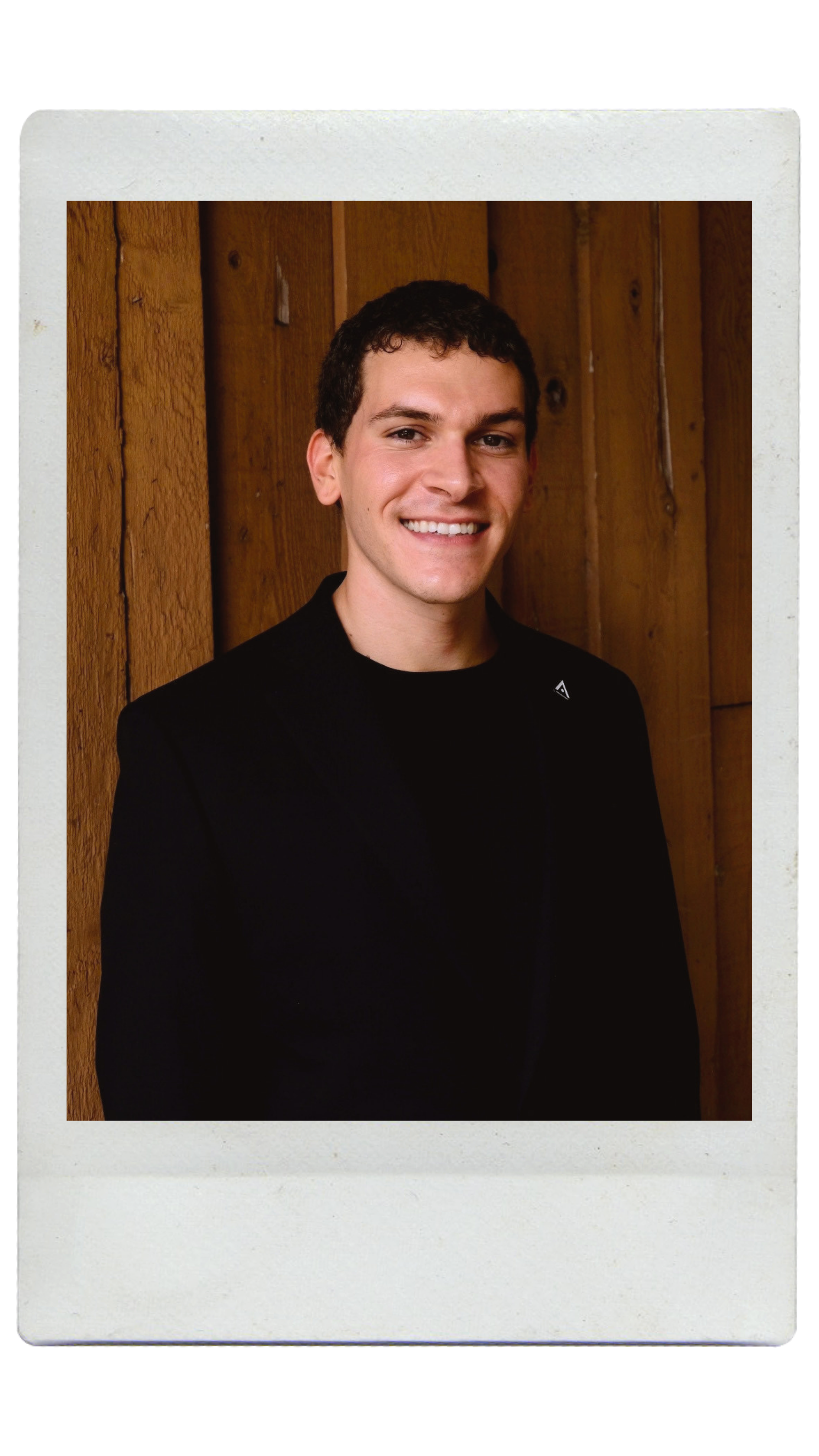 A young person with curly hair smiling at the camera, wearing a black shirt and layered necklaces, standing against a textured wall.