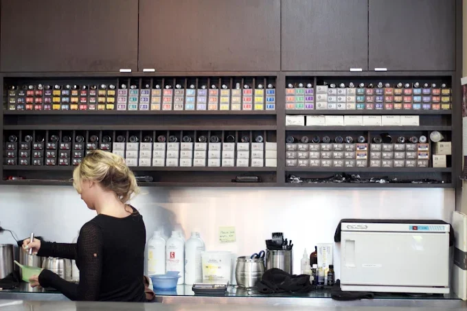 A woman preparing drinks or food in a kitchen, with a spice rack shelf above her filled with colorful spice jars and containers.