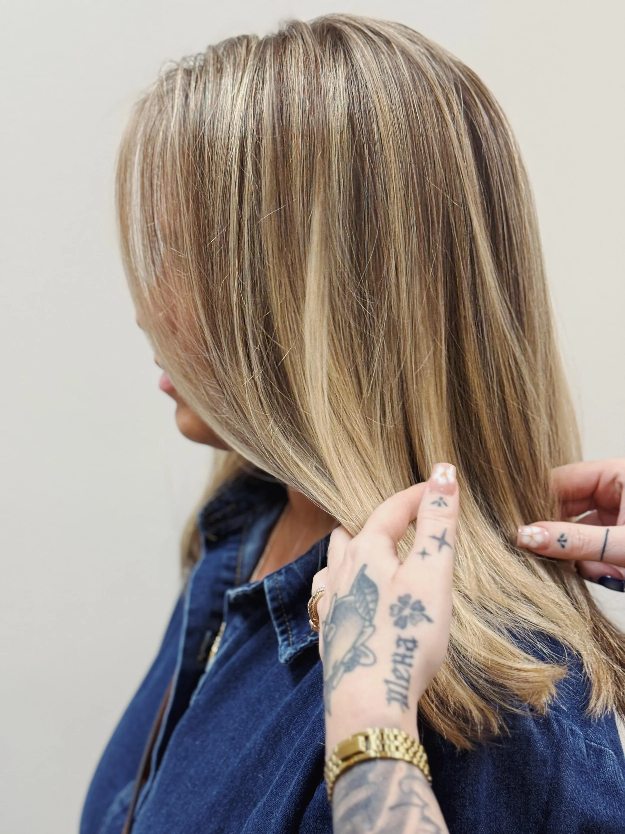 A woman with blonde hair in a bob cut, wearing a denim jacket, having her hair styled or checked by someone with tattooed hands.