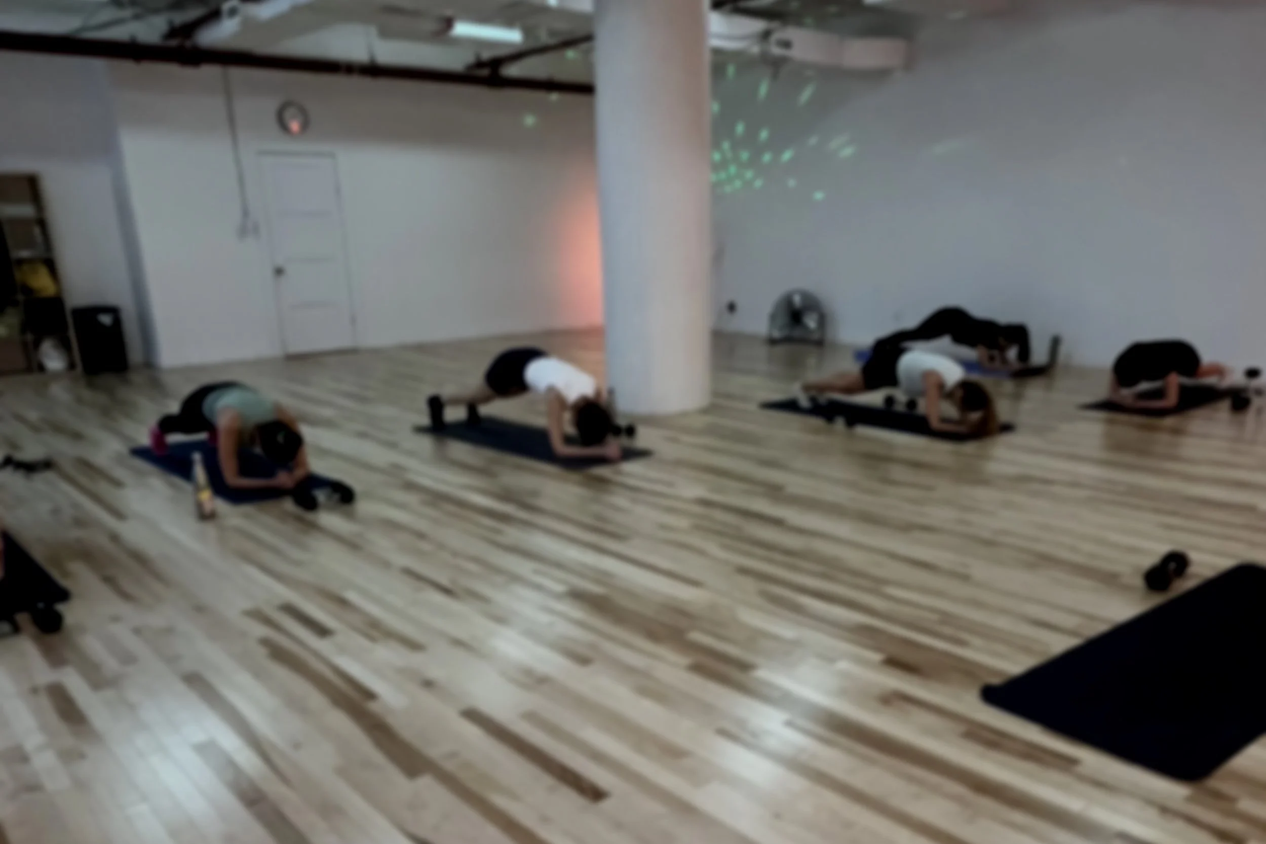 Four people doing yoga poses on mats in a studio with wooden floors and a white wall, a drum in the back corner, and a star-shaped light projection on the wall.