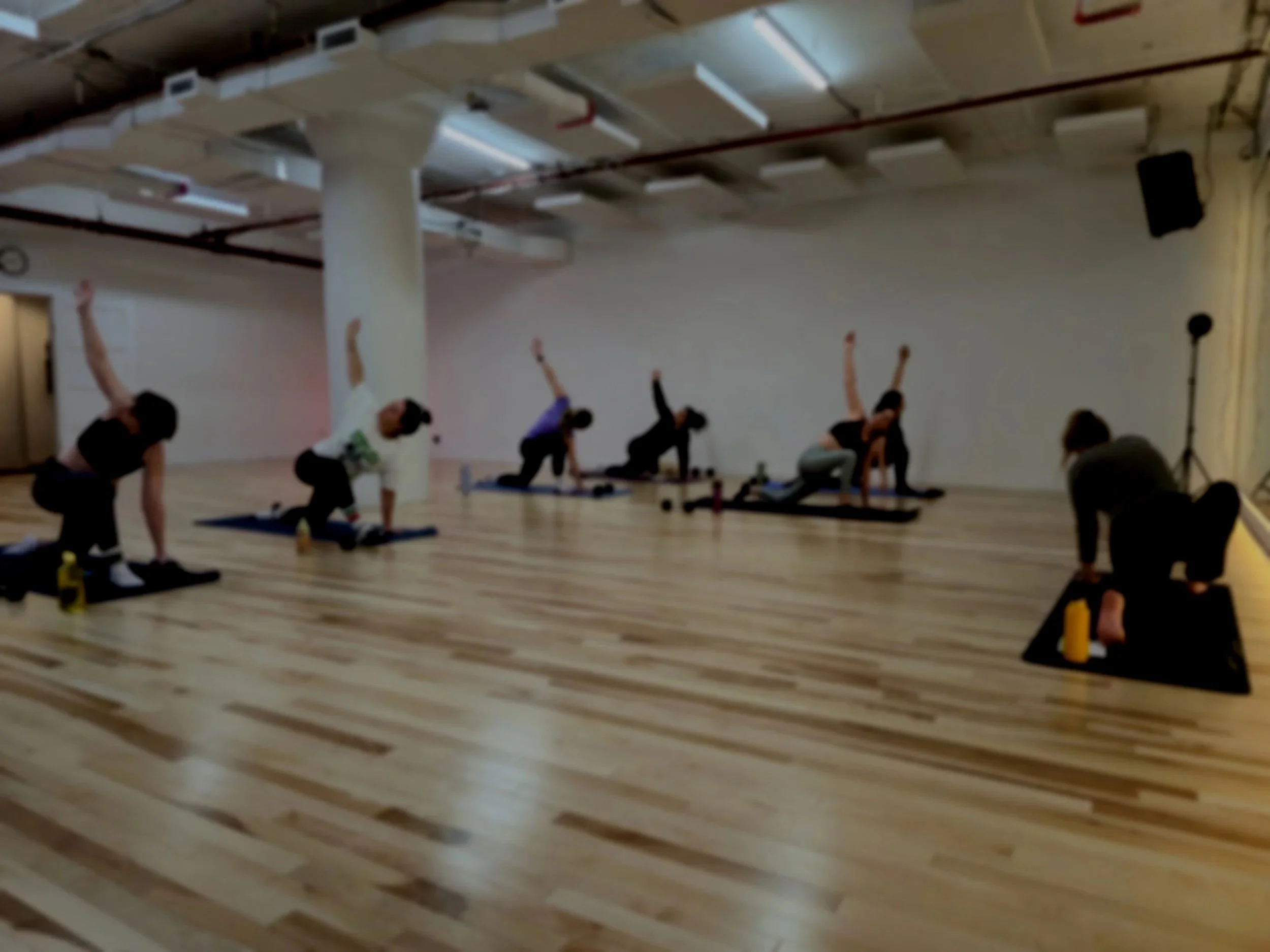 Group of people participating in a yoga or exercise class in a spacious room with wooden floors, some standing and some on mats, led by an instructor.