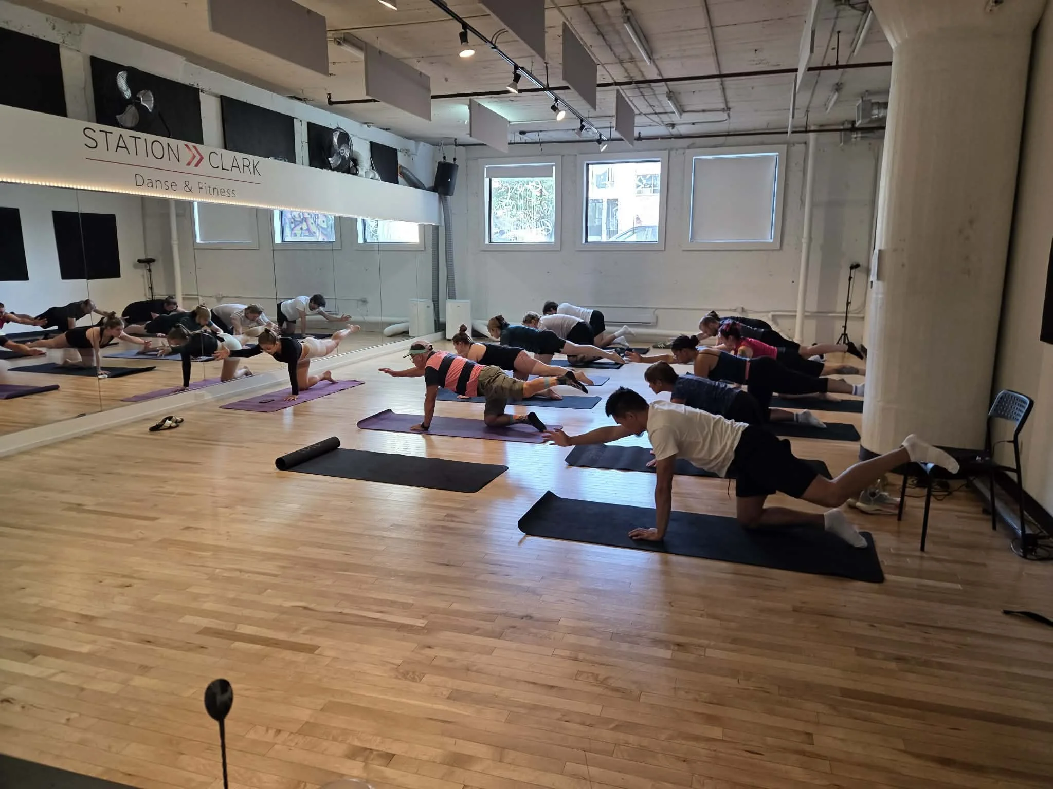 Group of people participating in a yoga or stretching class in a dance studio, performing an extended arm and leg pose on yoga mats, with ballet barre and mirrors on the wall.