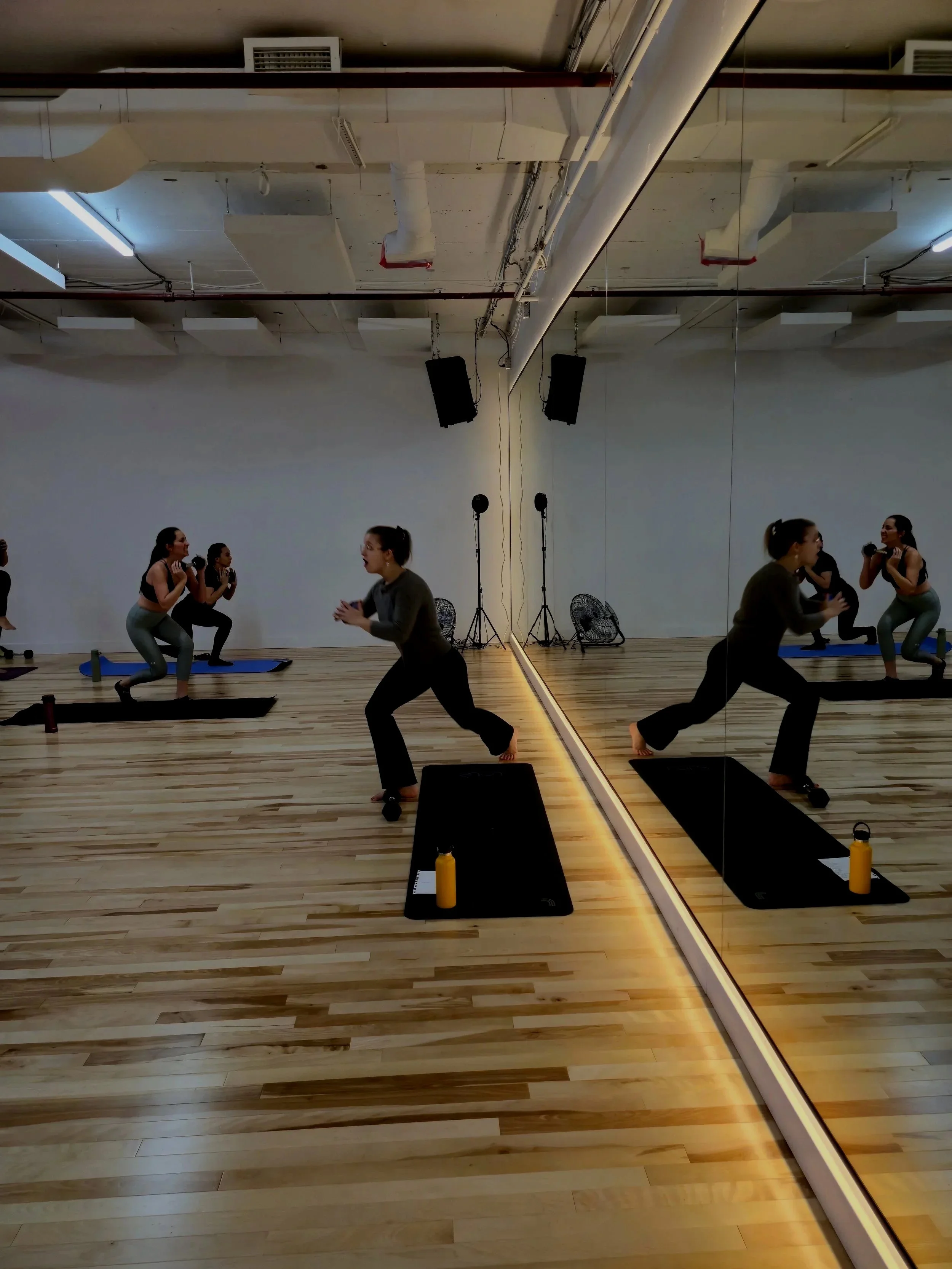 A group fitness class with women doing squats in a gym studio, with a large wall mirror and wooden flooring, two black speakers mounted on the ceiling, and water bottles on yoga mats.