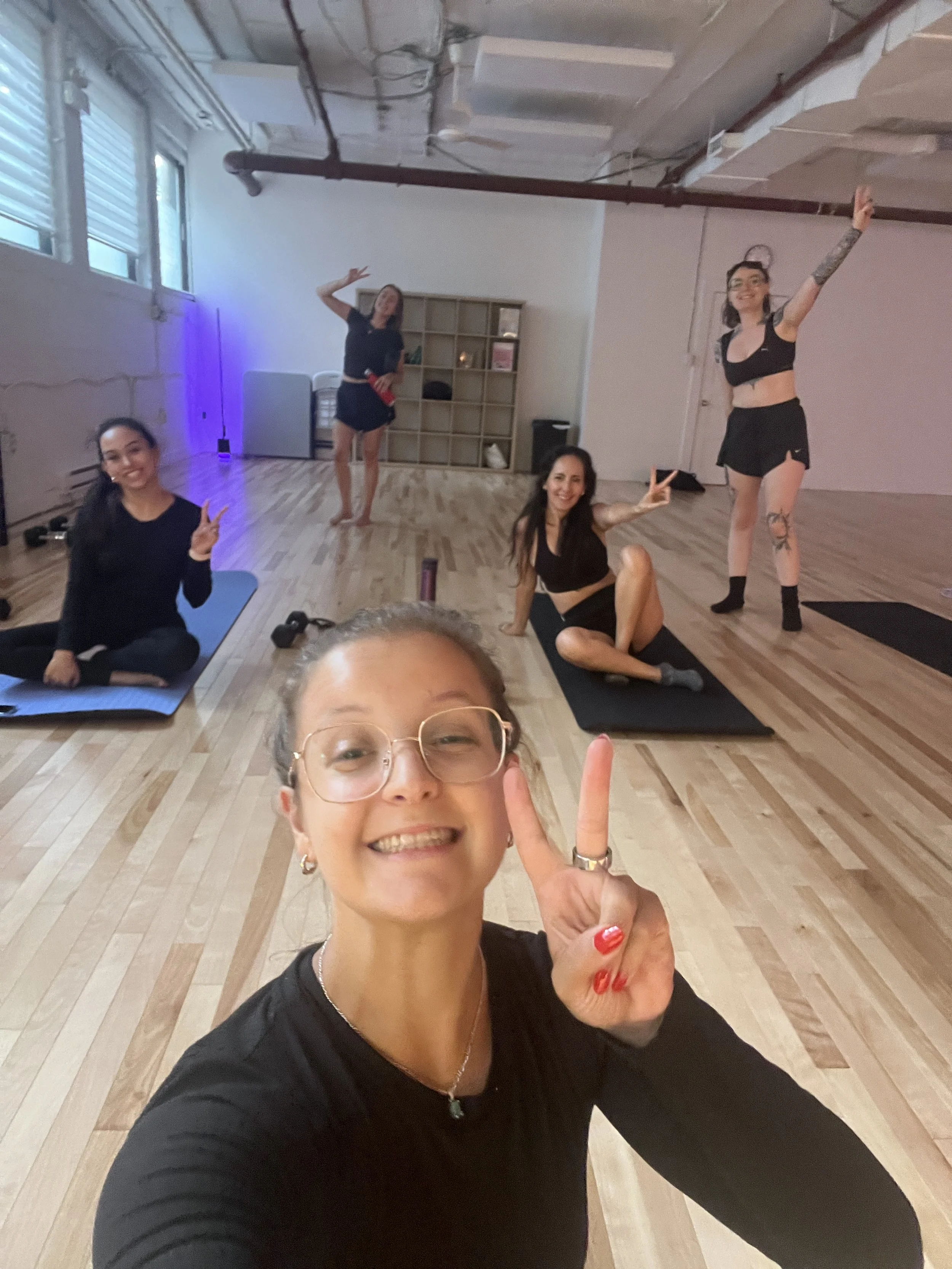 A group of women in workout clothes in a fitness studio, posing for a selfie; one woman in the foreground making a peace sign and smiling, others in the background making peace signs and waving.