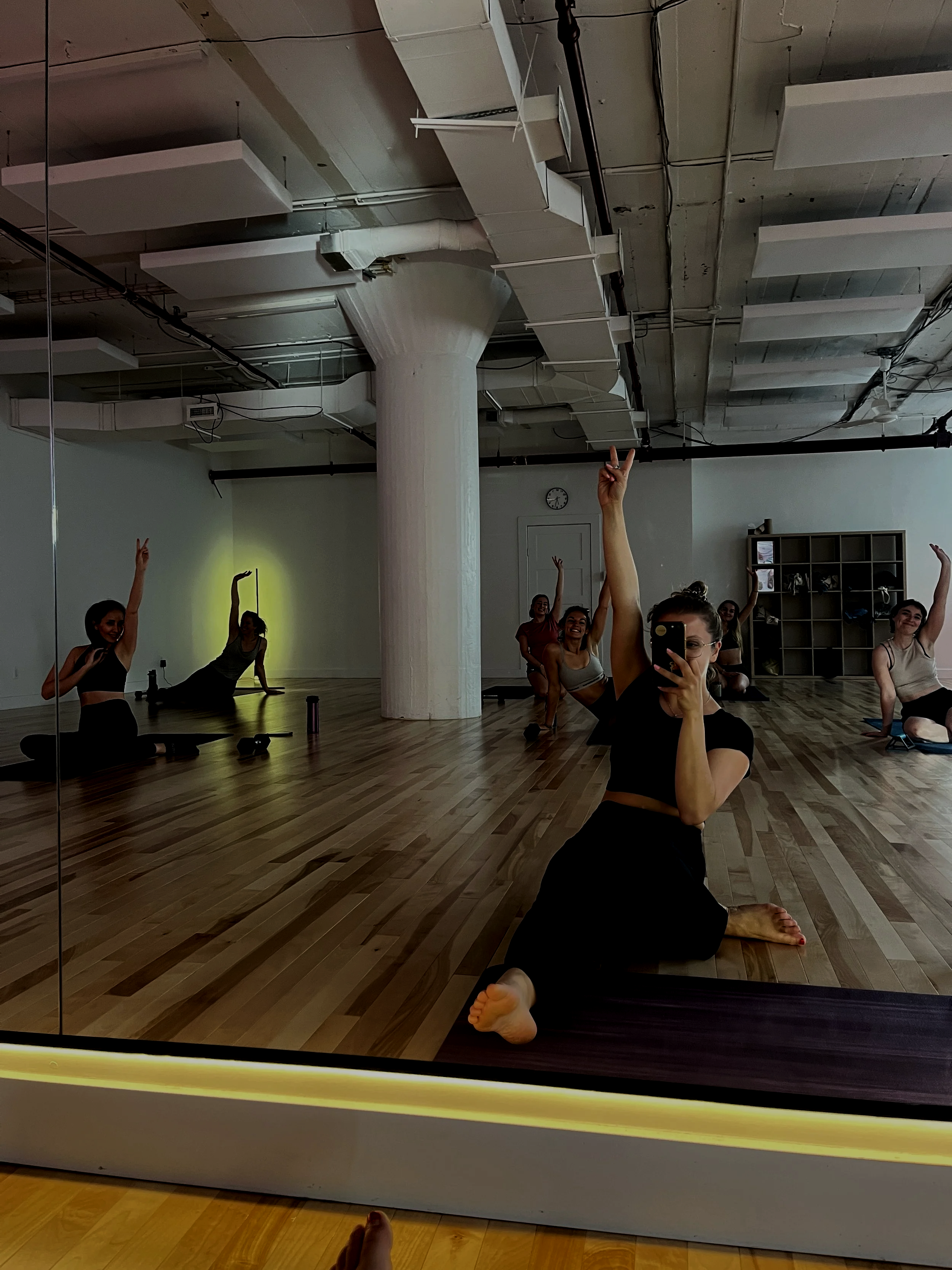 A group of people doing fitness exercises in a dance studio with a large mirror, wooden floor, white walls, exposed pipes, and a wall clock, some participants raising their arms, one taking a selfie, and others sitting on the floor.
