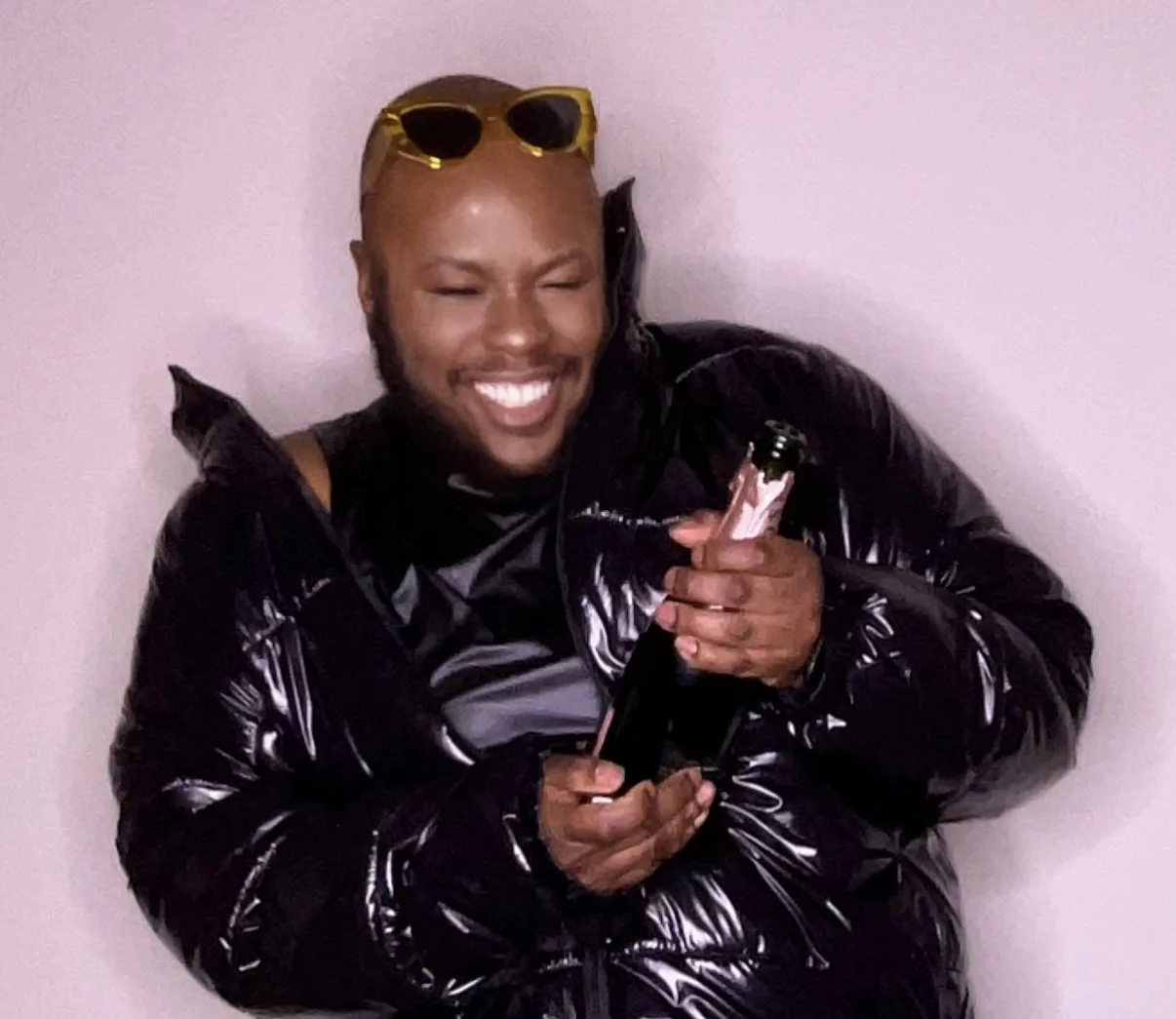 CARMINE DAVIS with sunglasses on their head, wearing a shiny black jacket, holding a bottle while standing against a plain wall.