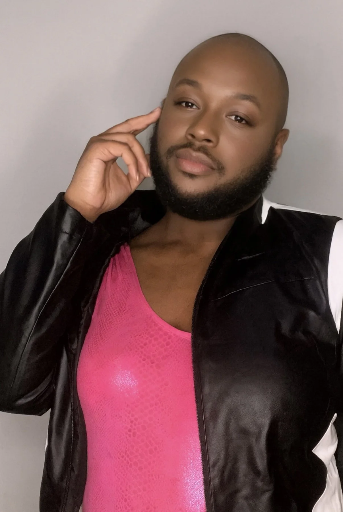Carmine Davis posing indoor against a plain light gray background. They are wearing a pink top and a black jacket with white accents, and are touching their face with their right hand while looking at the camera.