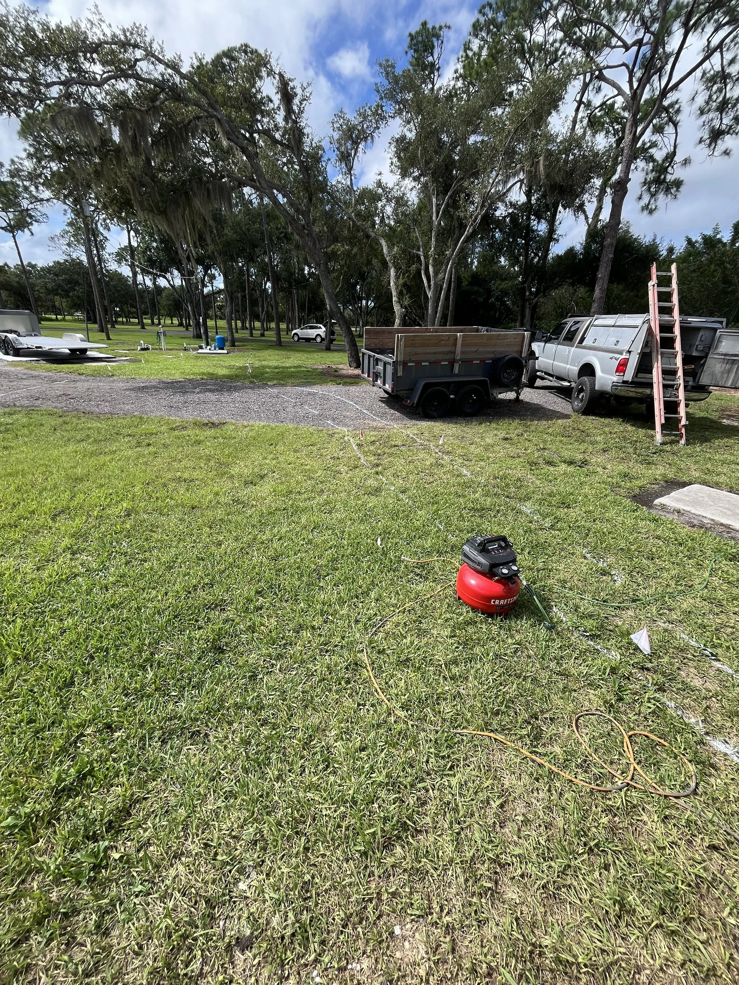 A grassy outdoor area with trees, a parked white pickup truck with a ladder on its back, a small trailer attached to the truck, and an equipment vacuum cleaner on the grass. There are several vehicles and equipment in the background.