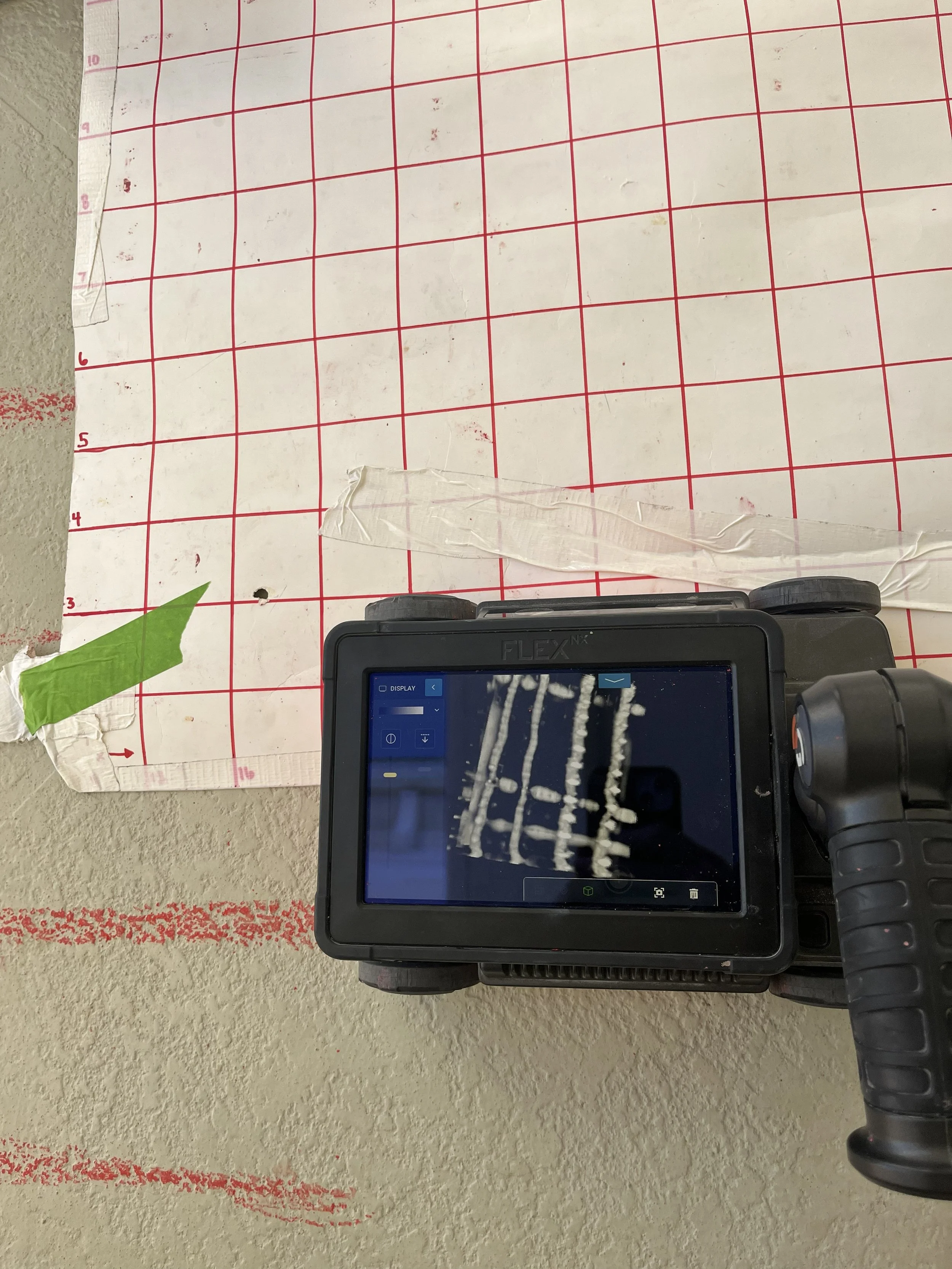 A meat slicing machine displaying sliced meat on the screen, placed on a textured surface with a white tiled grid paper and some green and clear tape nearby.