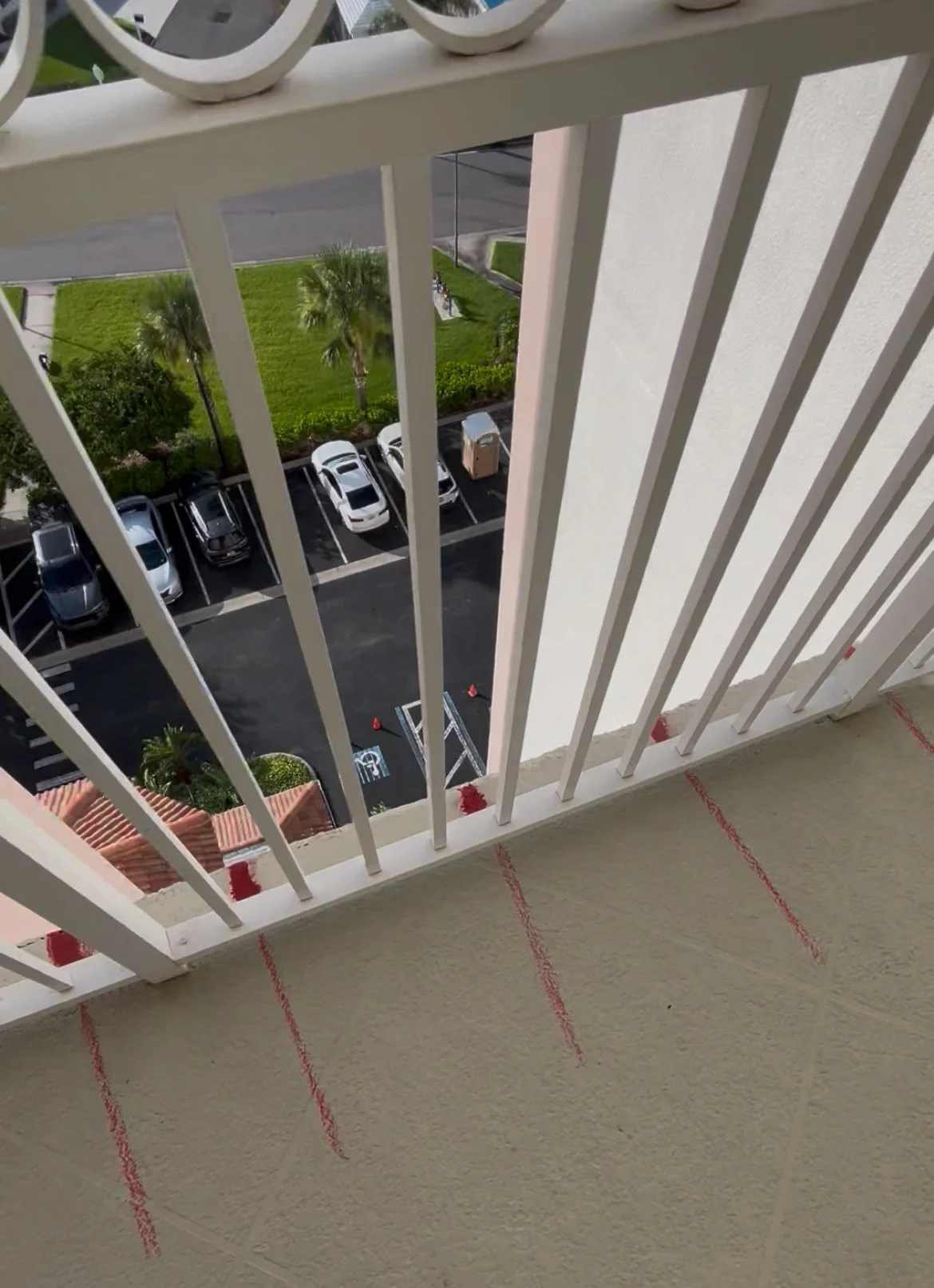View from a balcony overlooking a parking lot with several cars, green grass, palm trees, and a sidewalk beyond.