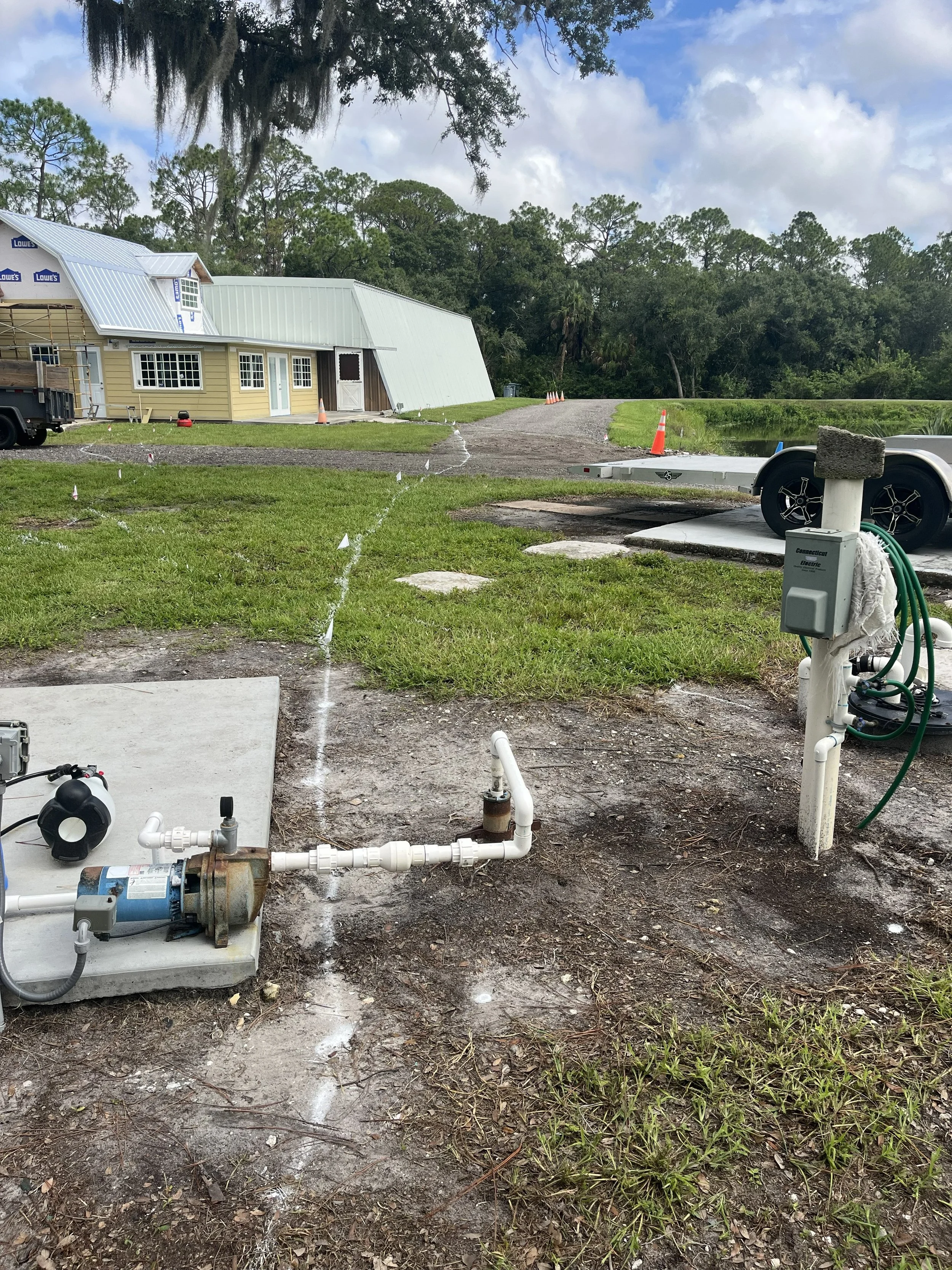 A construction site near a house with a metal roof, showing white pipes and electrical equipment in the foreground, and a dirt driveway with orange traffic cones, surrounded by grass, trees, and a pond.