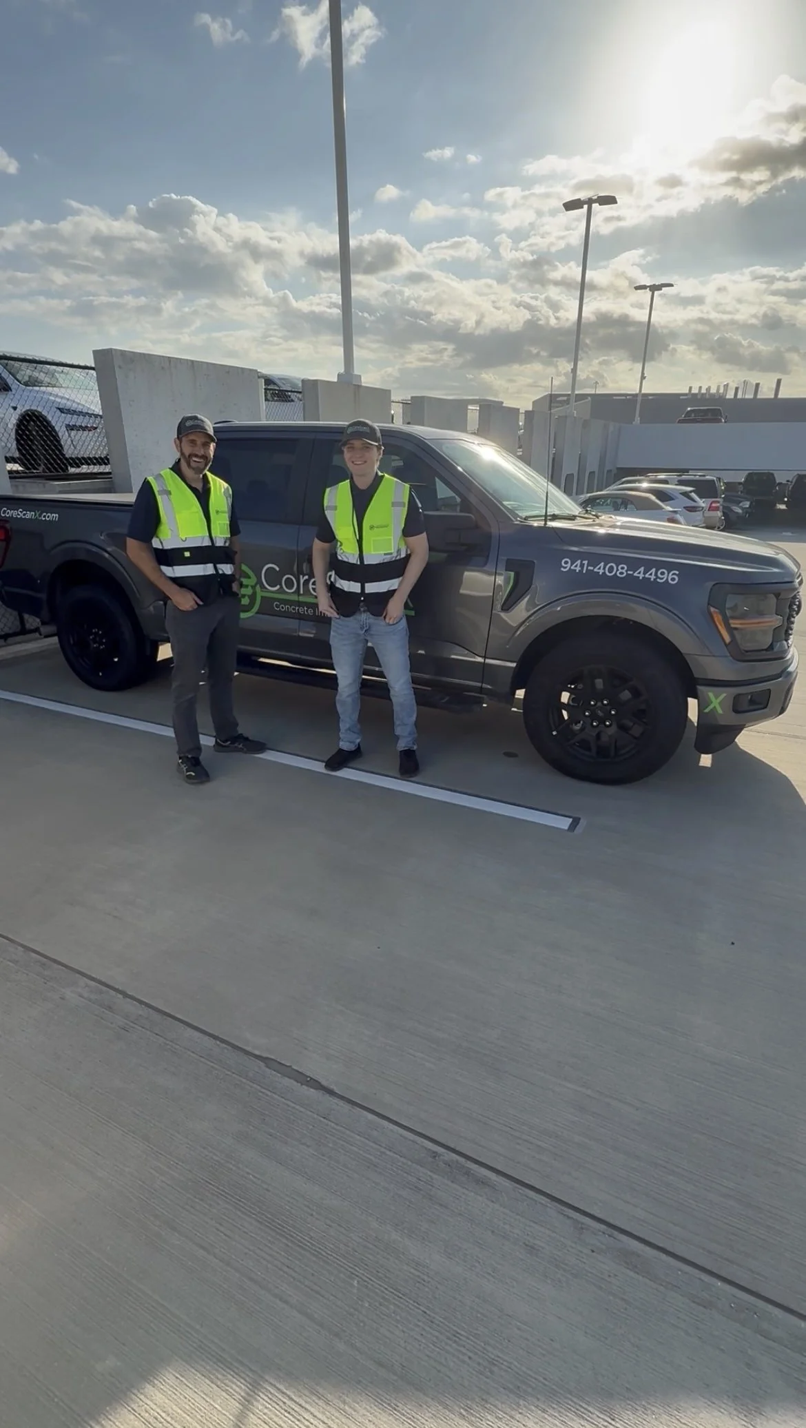 Two men wearing high-visibility vests standing next to a pickup truck with a company logo and phone number, in a parking lot during daytime with partly cloudy sky.