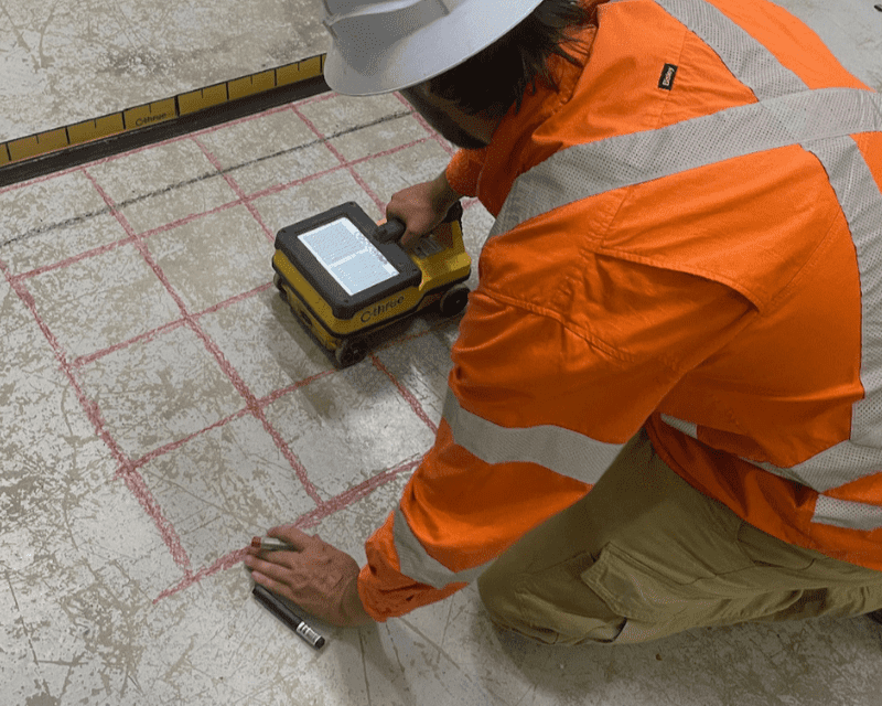 Construction worker in orange safety jacket and hard hat measuring concrete floor with a robotic device, marking a grid with chalk.