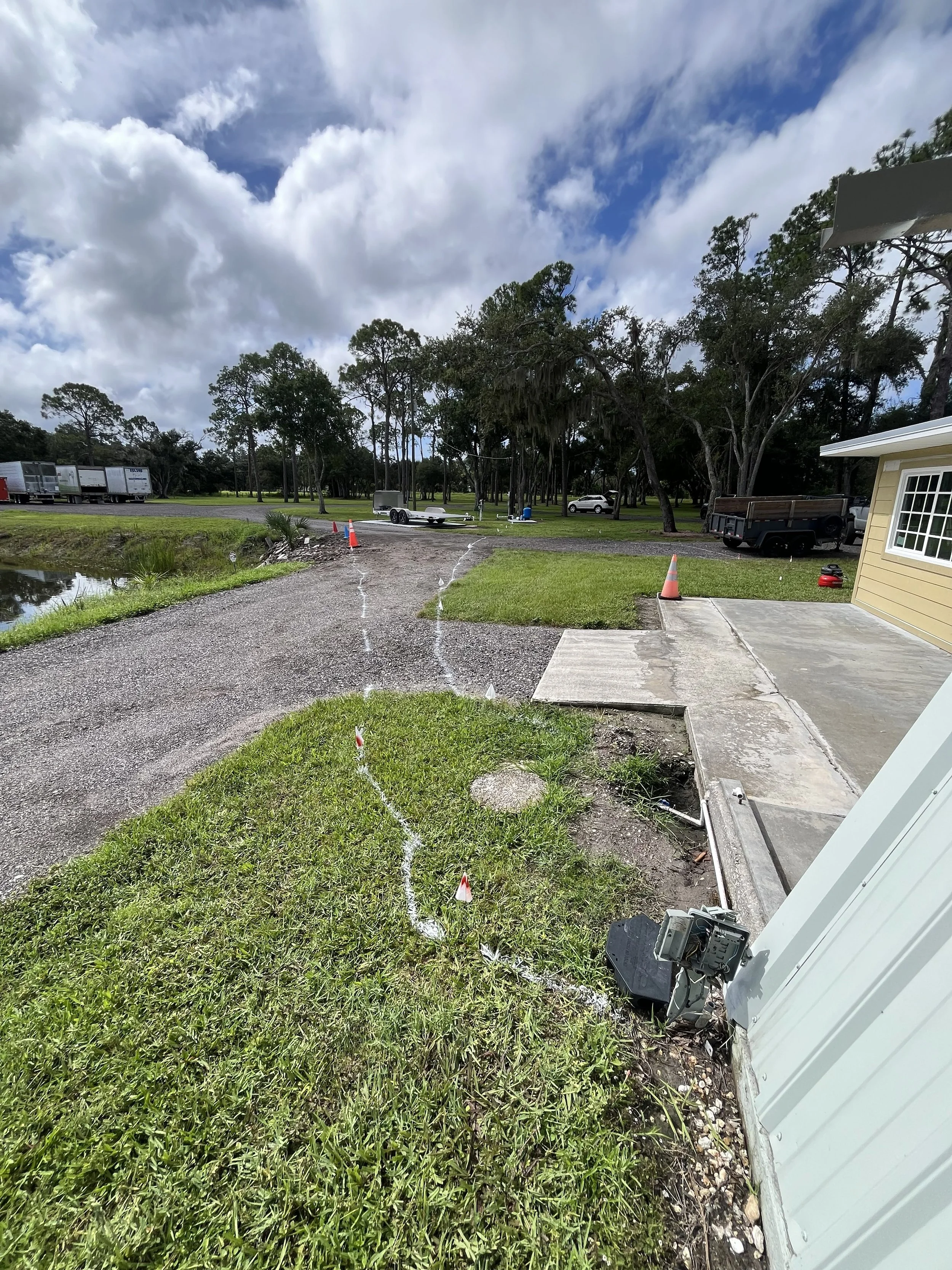A partially dug pathway with orange cones, a grassy area, a small body of water, trees, vehicles, trailer, and construction equipment under a cloudy sky.
