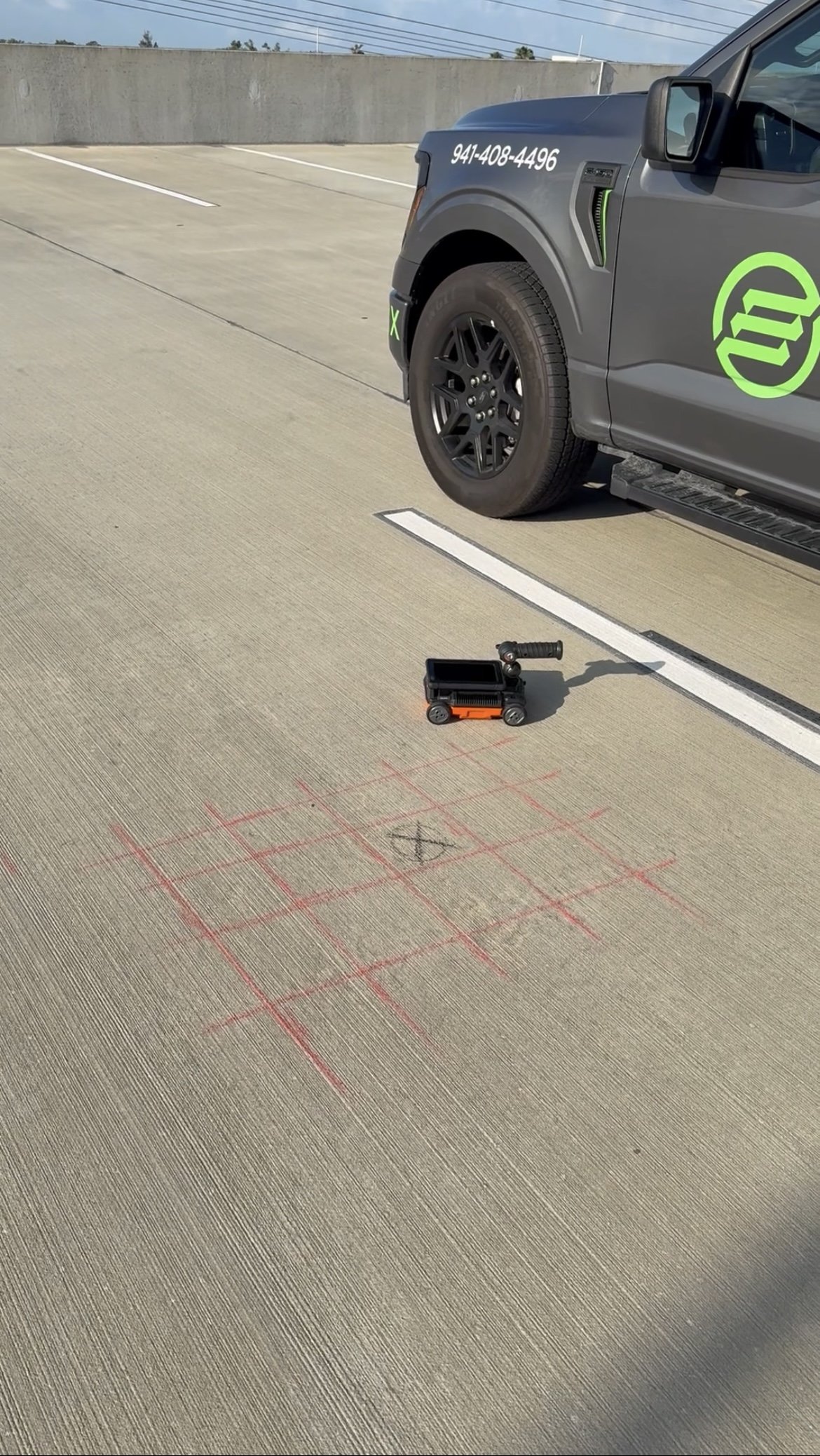 A parking lot with white lines and a concrete wall in the background. A gray truck with green and white logos, a phone number, and black wheels is parked on the right side of the image. On the ground near the truck, there are chalk markings and a small black cart.