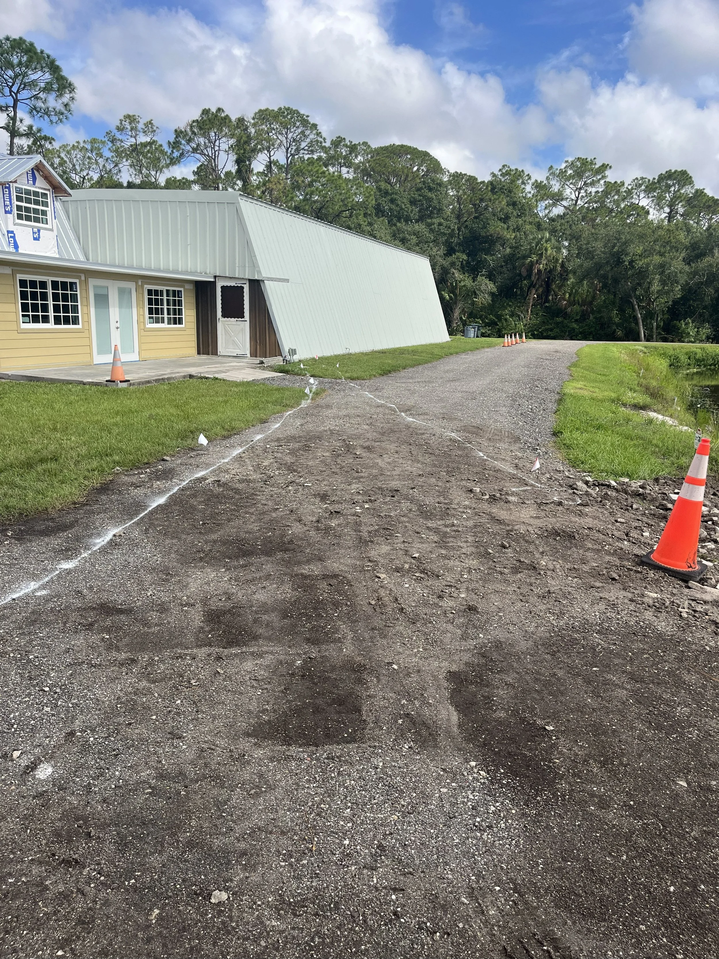 A gravel driveway leading to a small building with a yellow exterior and white doors, surrounded by green grass and trees, with construction cones placed along the side.