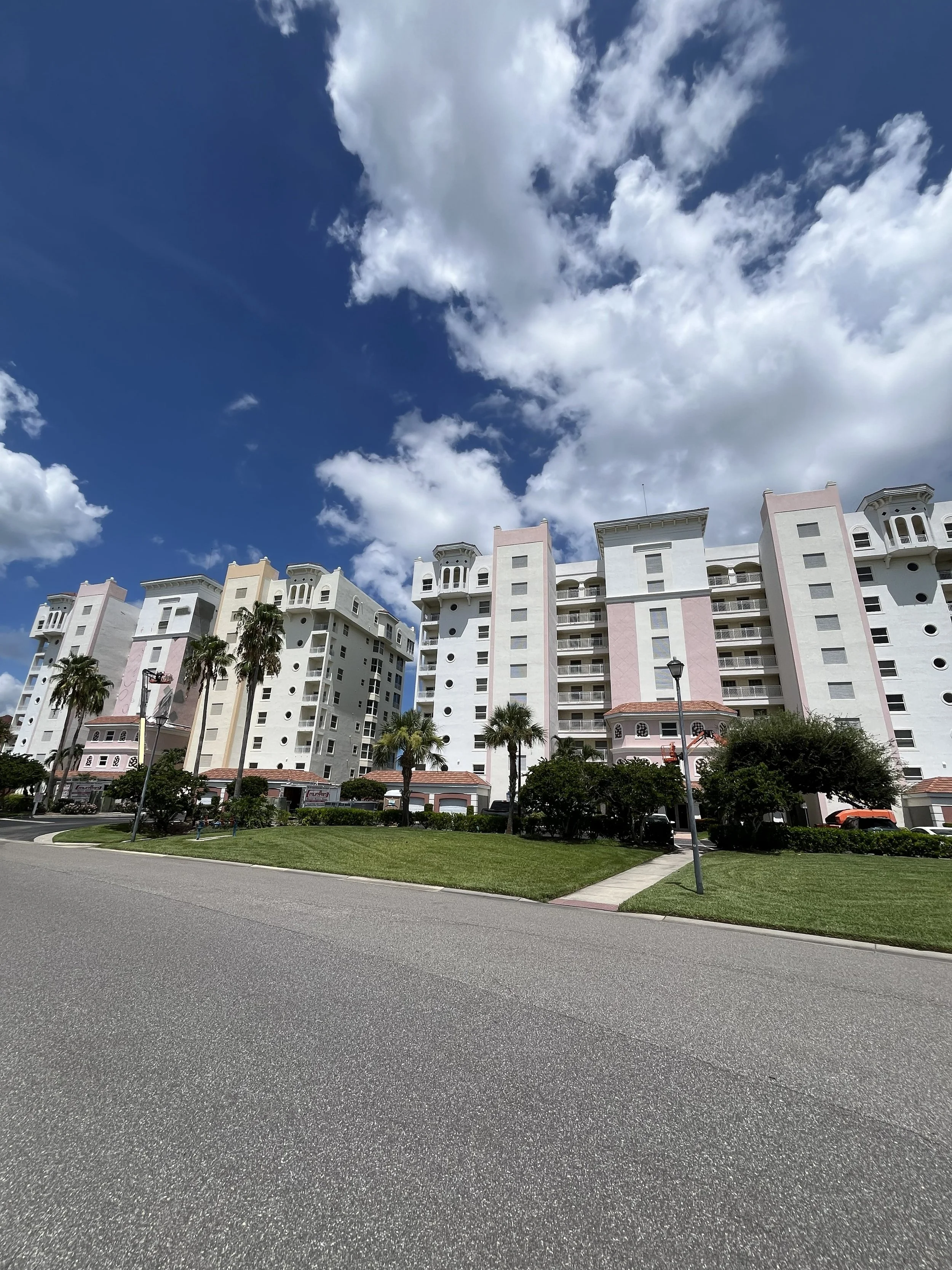 Apartment buildings with palm trees in front, green lawn, sidewalk, street, and partly cloudy blue sky.