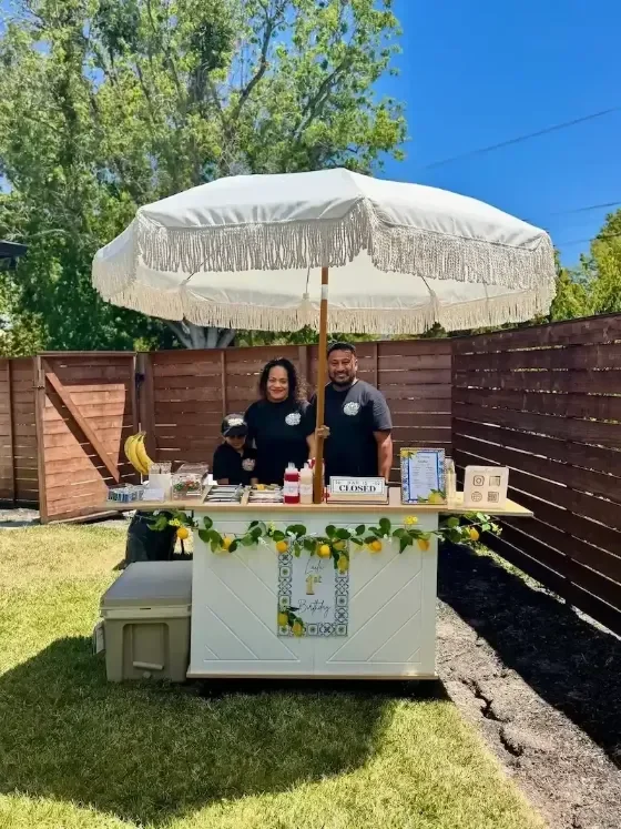Family serving at a white lemonade cart with a fringe umbrella