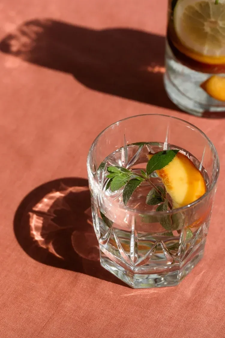 A glass of infused water with lemon, mint, and a slice of peach on a pink surface, casting a shadow.