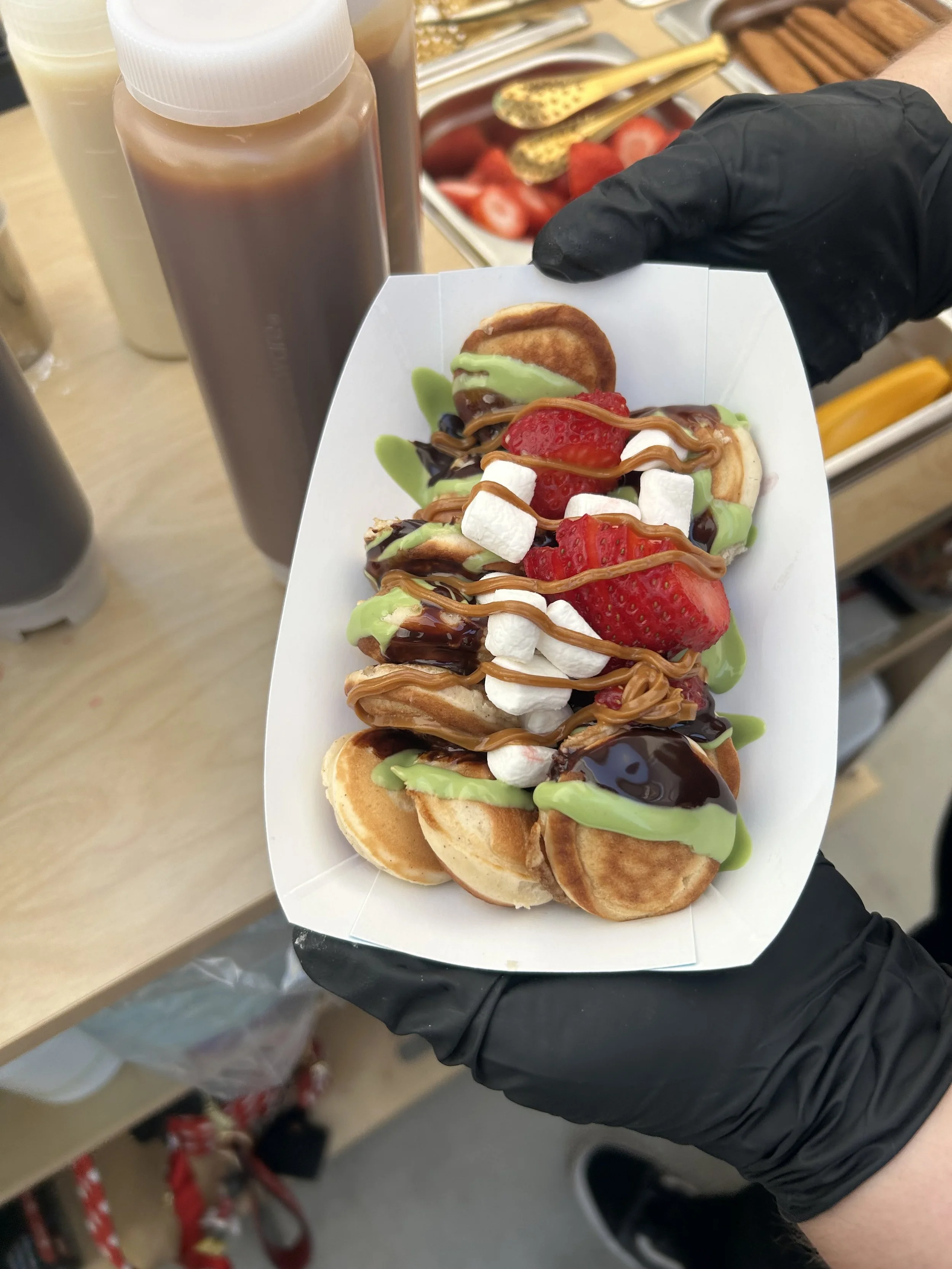A person wearing black gloves holding a tray of mini waffles topped with strawberries, marshmallows, chocolate syrup, and green sauce.