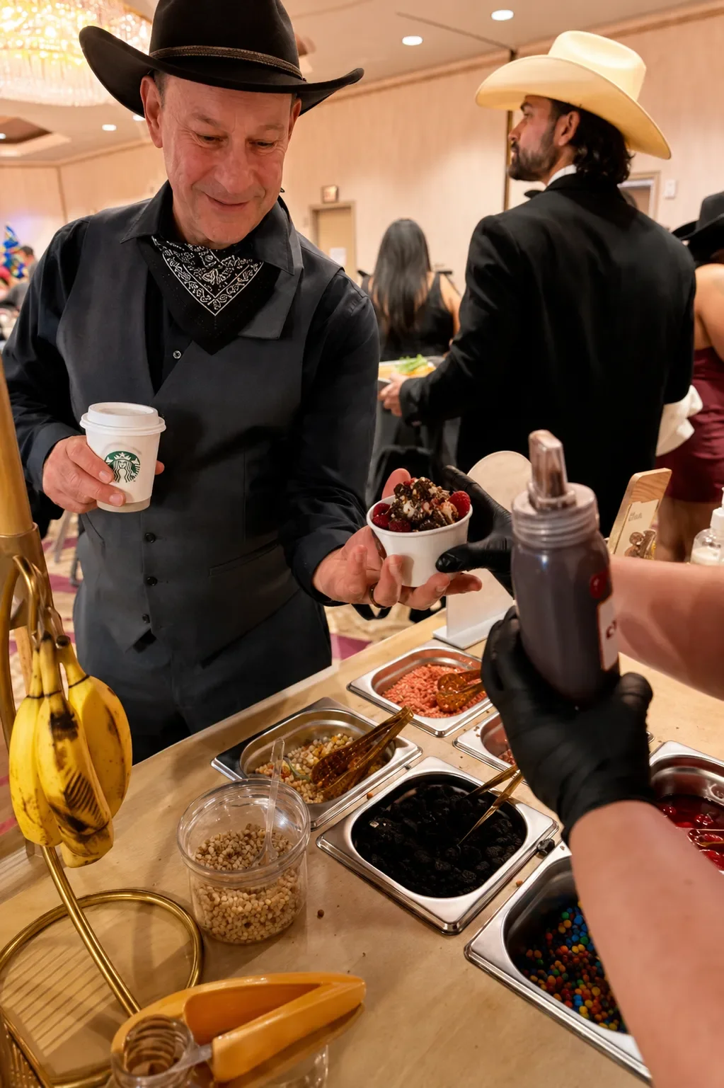 Man in cowboy hat receives a bowl of mixed berries and chocolate from server at dessert station, with toppings and desserts laid out on the counter, and other guests in the background.