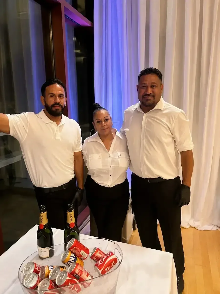 Three people dressed in white shirts and black pants standing together at a social event, with a table in front holding two bottles of champagne and a bowl of soda cans. The background includes curtains and glass windows.