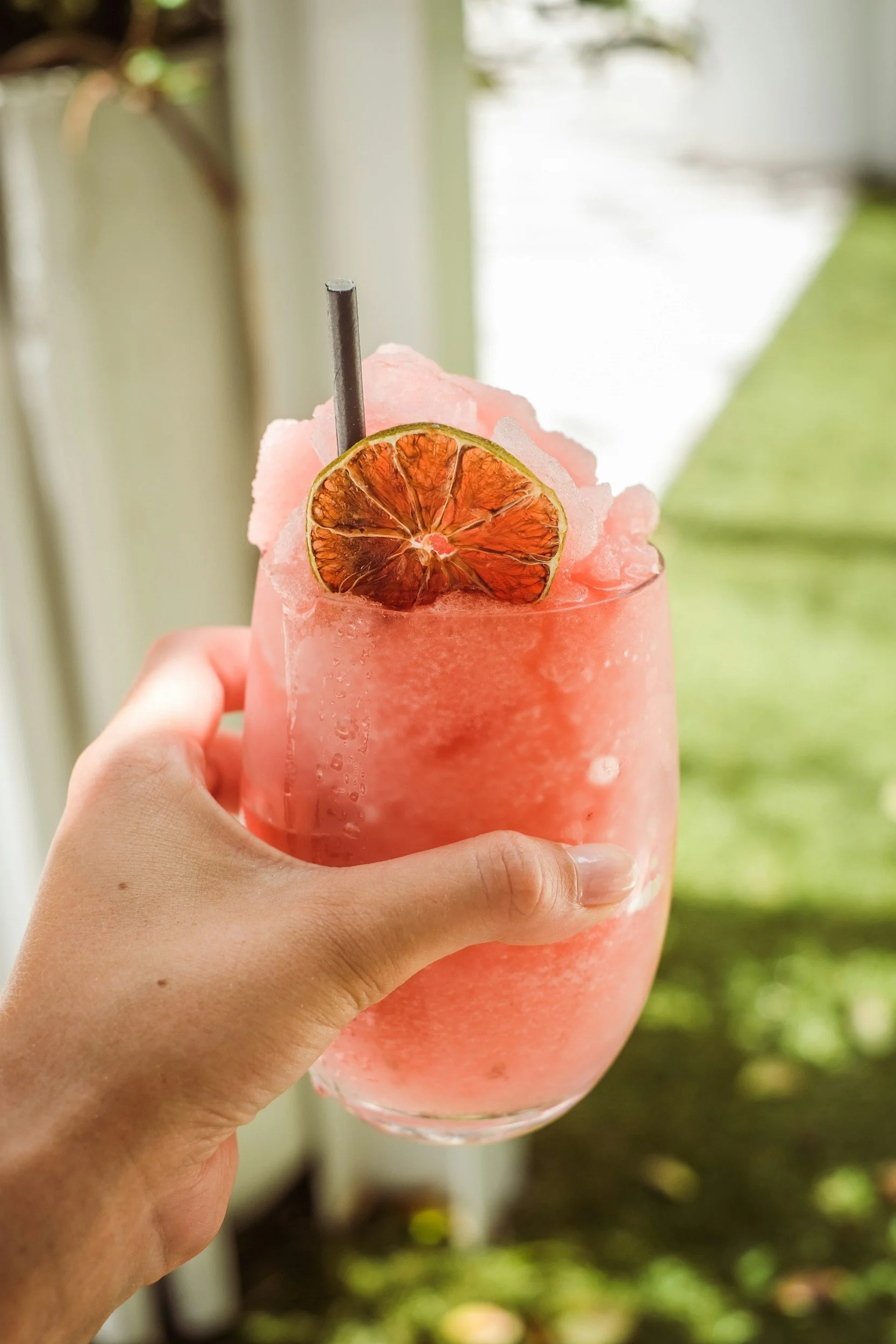A hand holding a tall glass of pink frozen beverage garnished with dried blood orange slices and a black straw, with a blurred outdoor background.